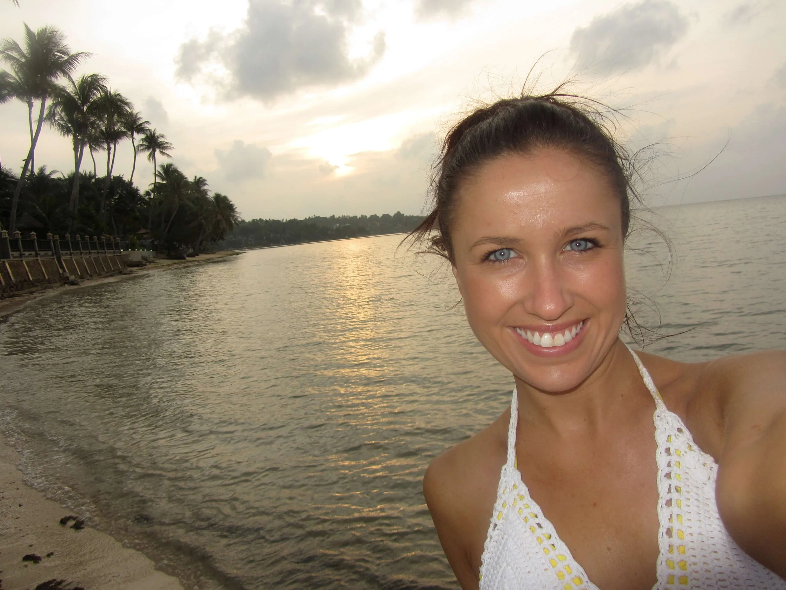 A woman smiling at the camera with wet hair, wearing a white crochet top, at the beach during sunset or sunrise, with palm trees and cloudy sky in the background.