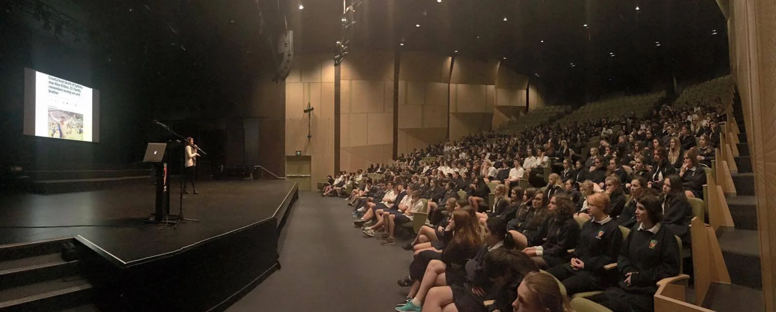 A large auditorium filled with students sitting in woven chairs, watching a presentation on stage where a woman is speaking into a microphone and a large screen displays a slide.