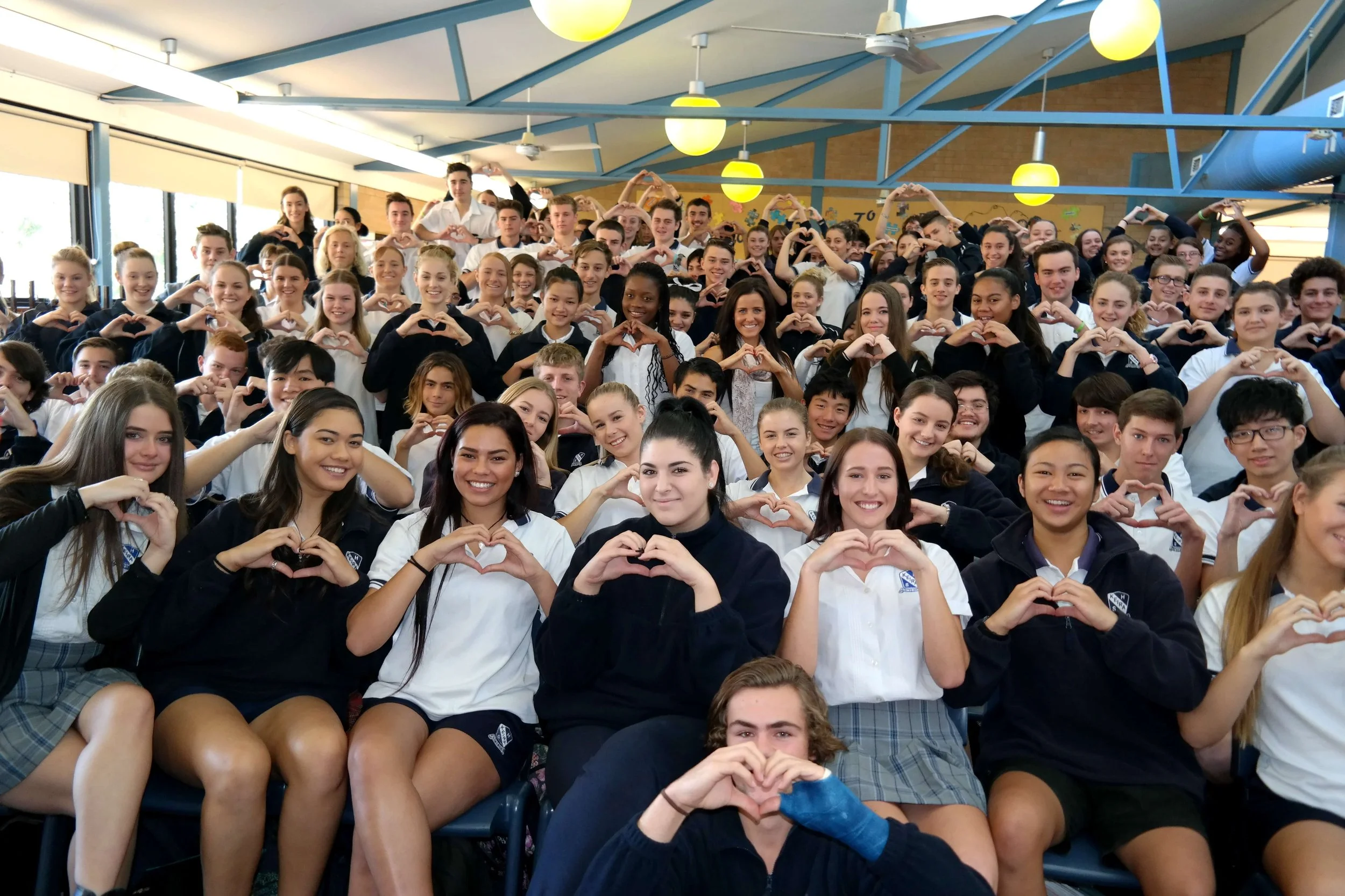 Group of students in school uniform inside a classroom, many of them making heart shapes with their hands.