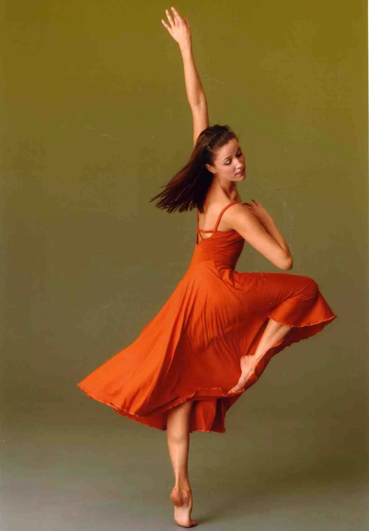 Dancer in an orange dress performing a pose, standing on one foot with the other leg lifted and bent, against a plain background.