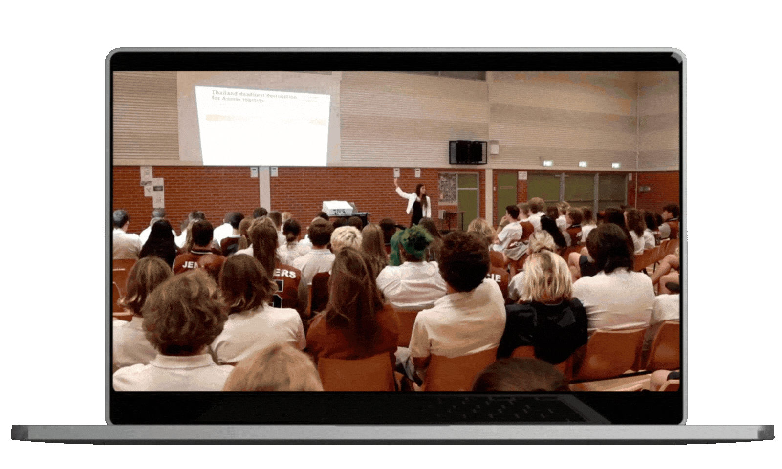 A classroom or conference room with a large group of students or attendees watching a woman giving a presentation. The woman is standing at the front near a projector and a screen, speaking and gesturing with her right hand. The room has brown brick walls and some doors on the side.