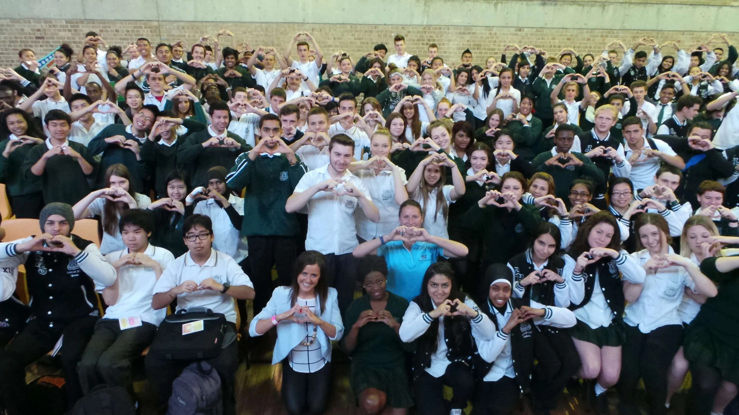 A large group of diverse students and teachers posing together in a school auditorium, making heart shapes with their hands.