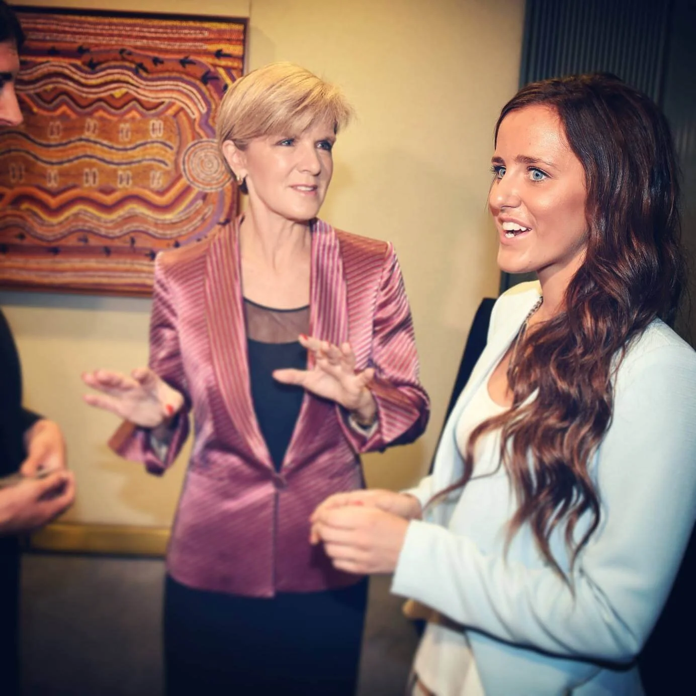 Two women having a conversation indoors, with decorative artwork on the wall behind them.