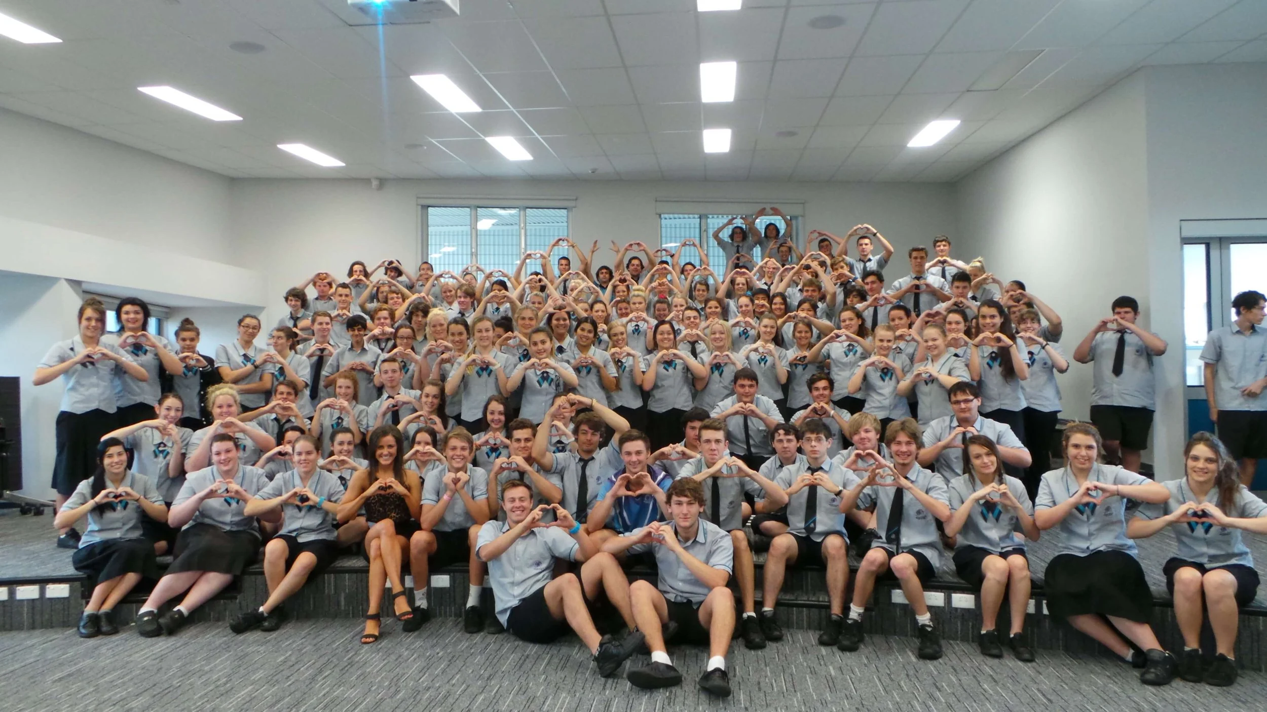 A large group of students in school uniforms gathered together in a classroom or auditorium, forming heart shapes with their hands.