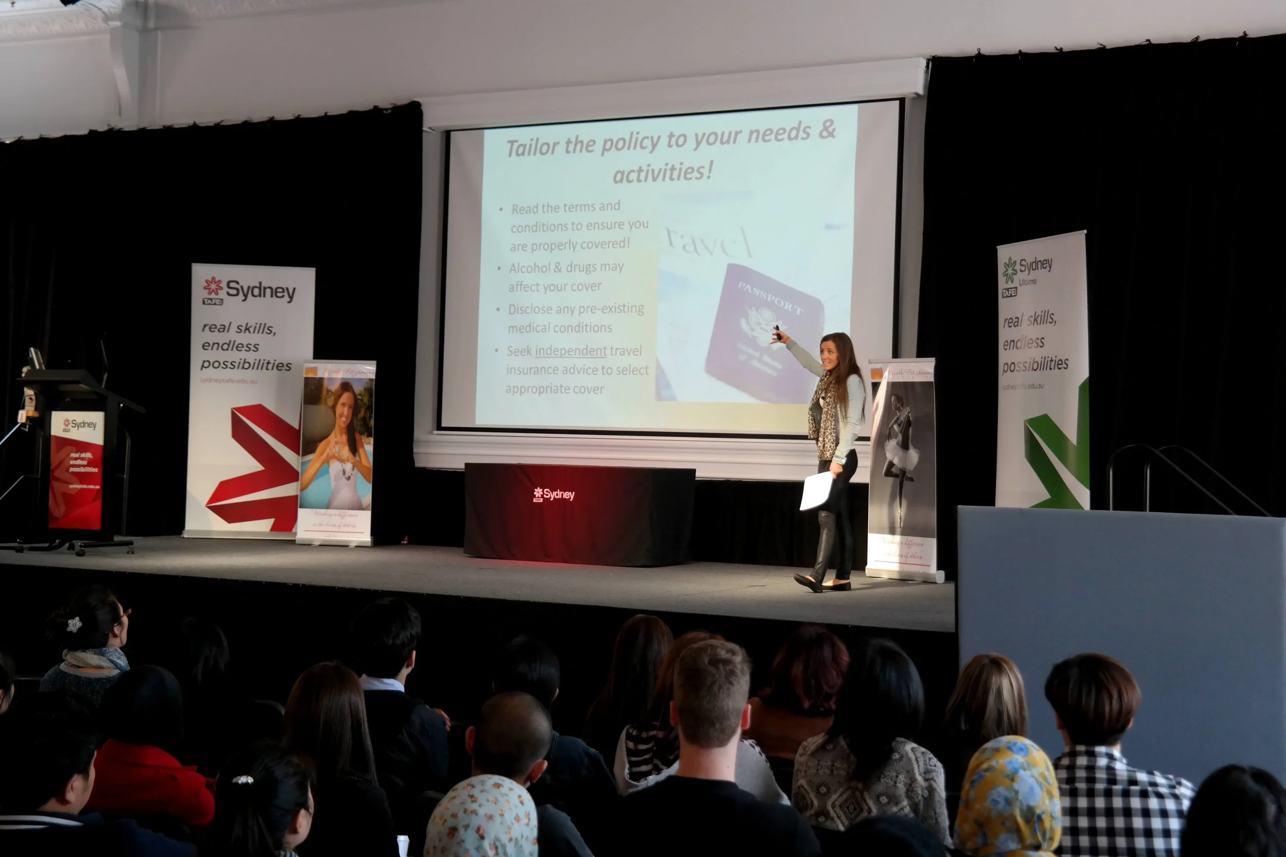 A woman giving a presentation on a stage at an event, with a projector screen behind her displaying a slide titled 'Tailor the policy to your needs & activities!' There are banners on either side of the stage with the Sydney TAFE logo and slogans.