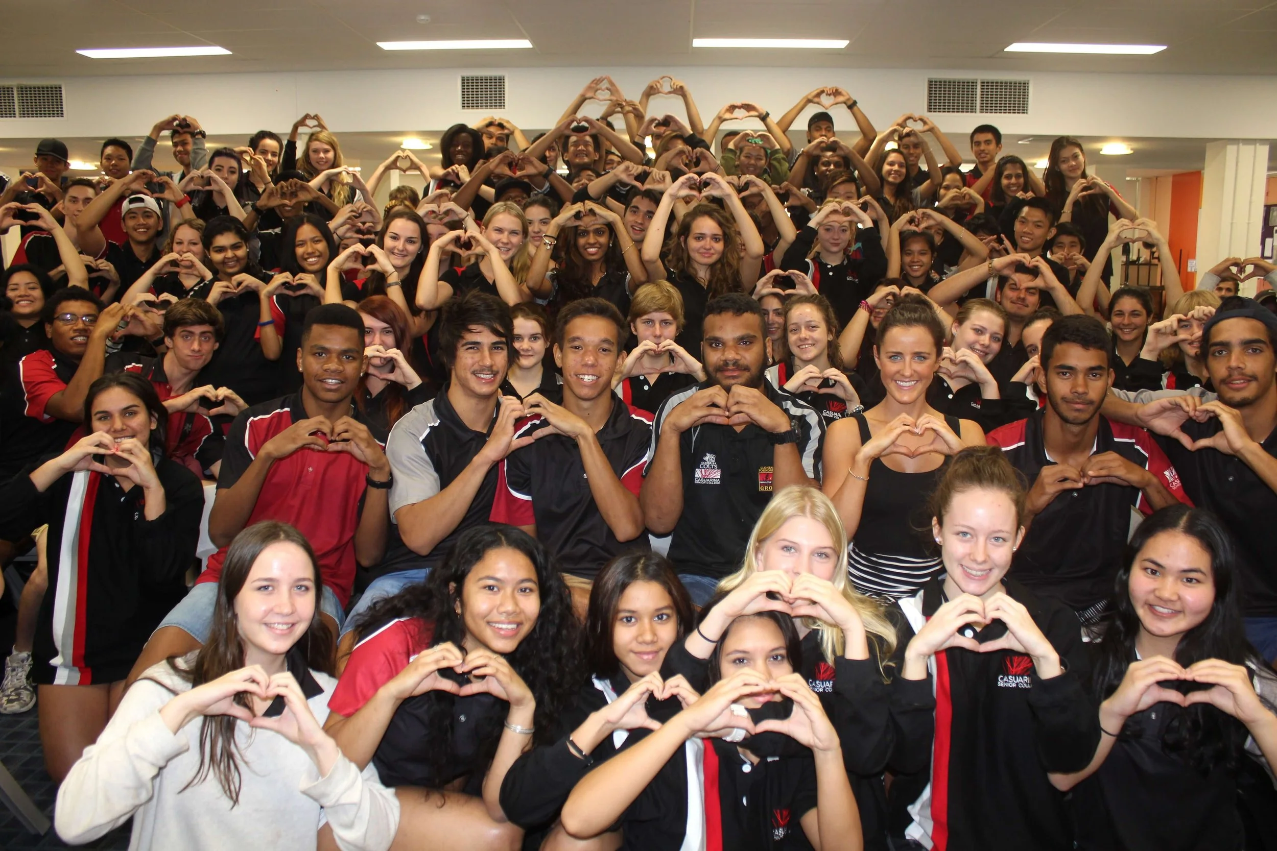 A large group of diverse young students indoors, making heart shapes with their hands, smiling and looking at the camera.