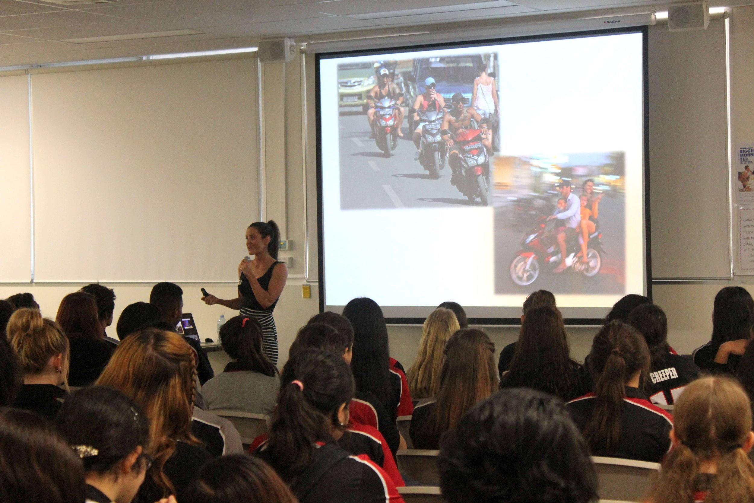 A woman giving a presentation to a large audience in a classroom or conference room, with a large screen behind her displaying a photo of multiple motorbikes and people, taken during daytime on a busy street.