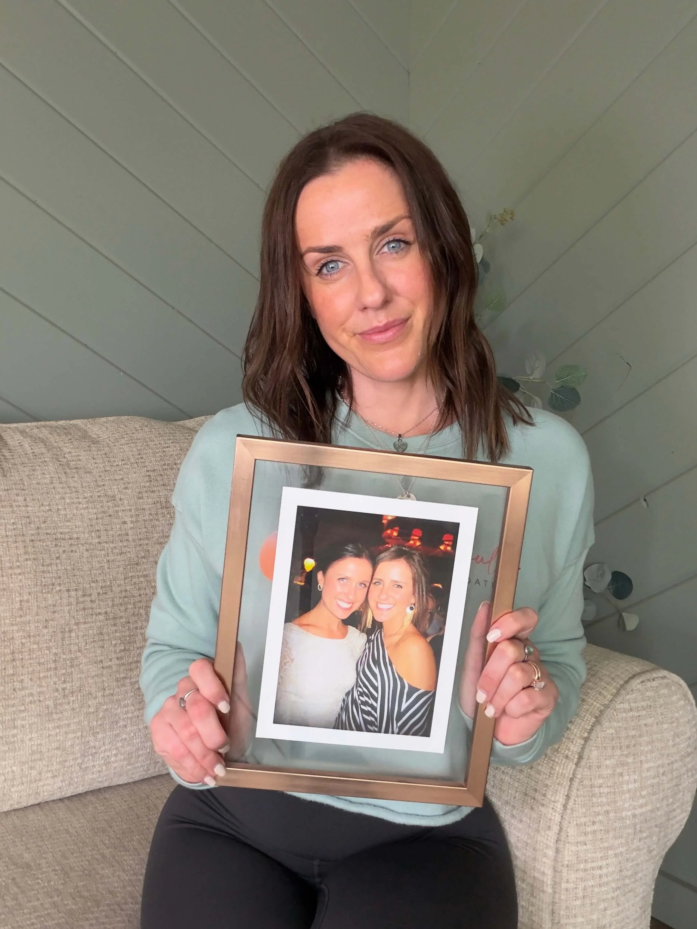 A woman with blue eyes and brown hair is sitting on a beige couch, holding a framed photograph of two women smiling and posing together at a social event.