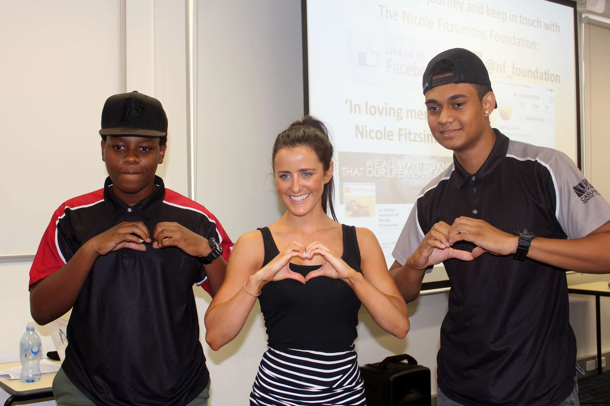 Three smiling people standing close together in a classroom, making heart shapes with their hands. Two young men are on the sides, and a woman is in the middle. A presentation screen with text is visible in the background.
