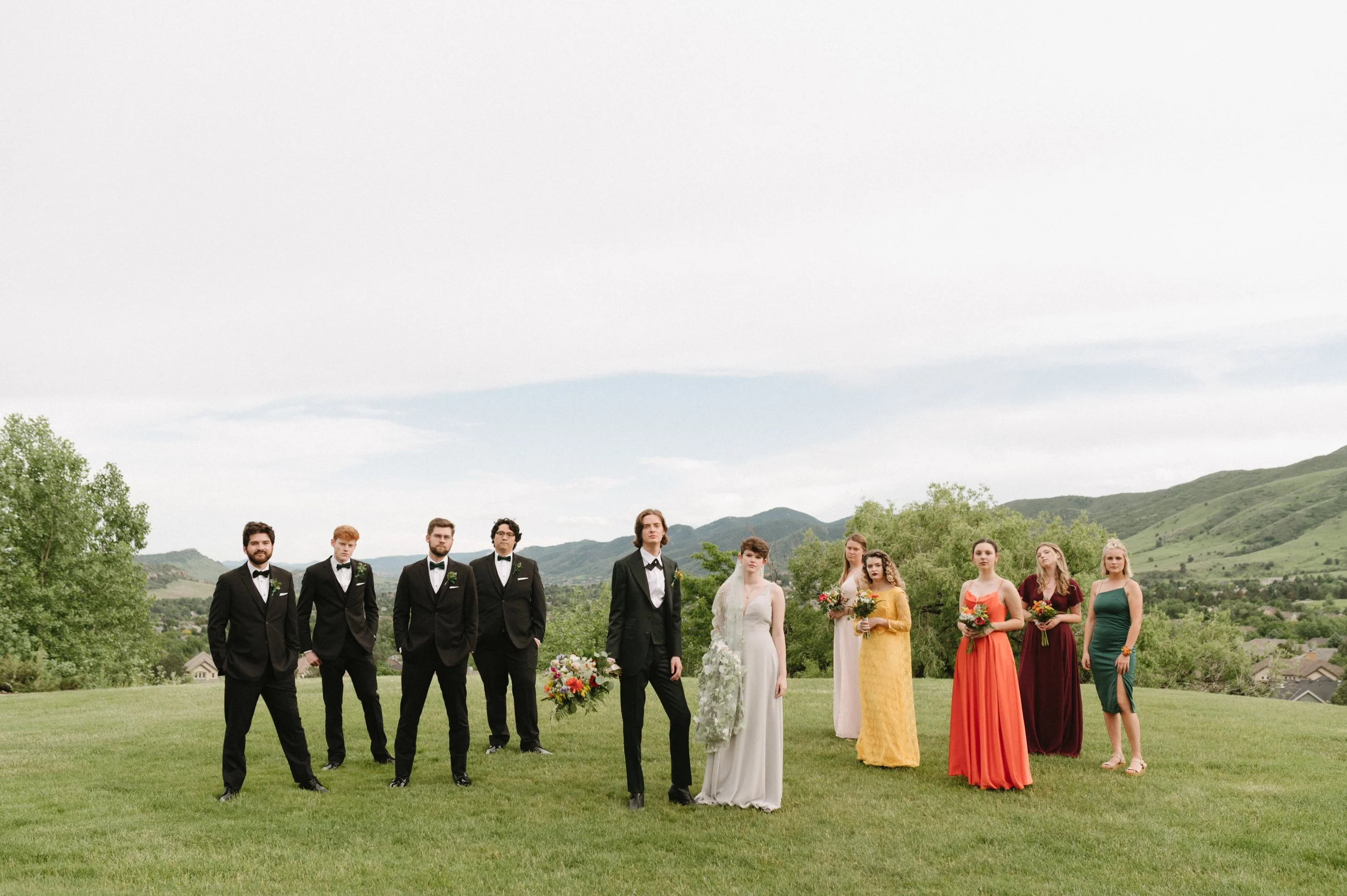 A wedding party standing on a grassy field with rolling hills and trees in the background. The group includes five men in black tuxedos, the bride in a white dress, and four women in colorful dresses holding bouquets.