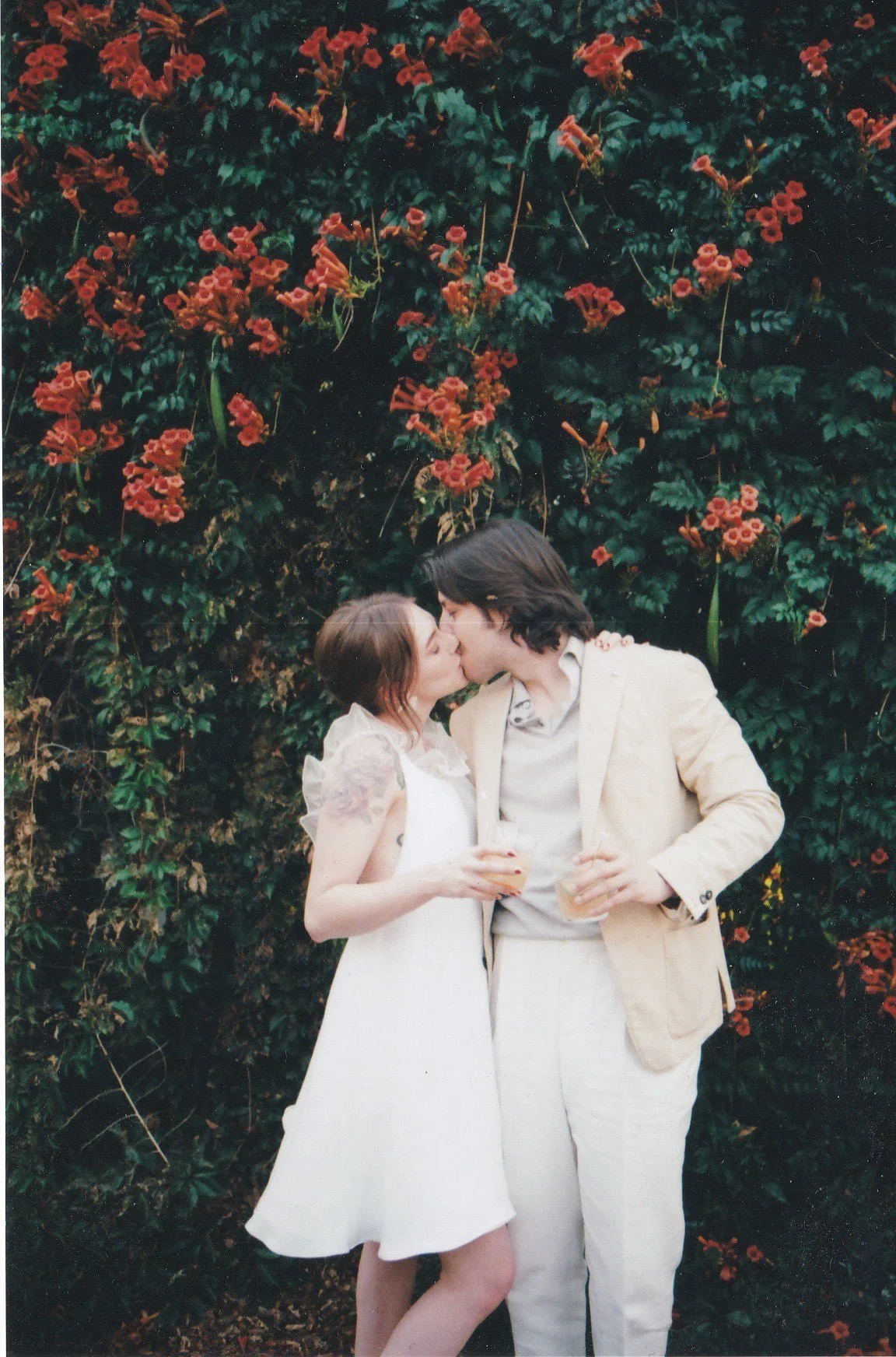 A bride and groom sharing a kiss at their backyard wedding, with a backdrop of blooming red flowers and green bushes.