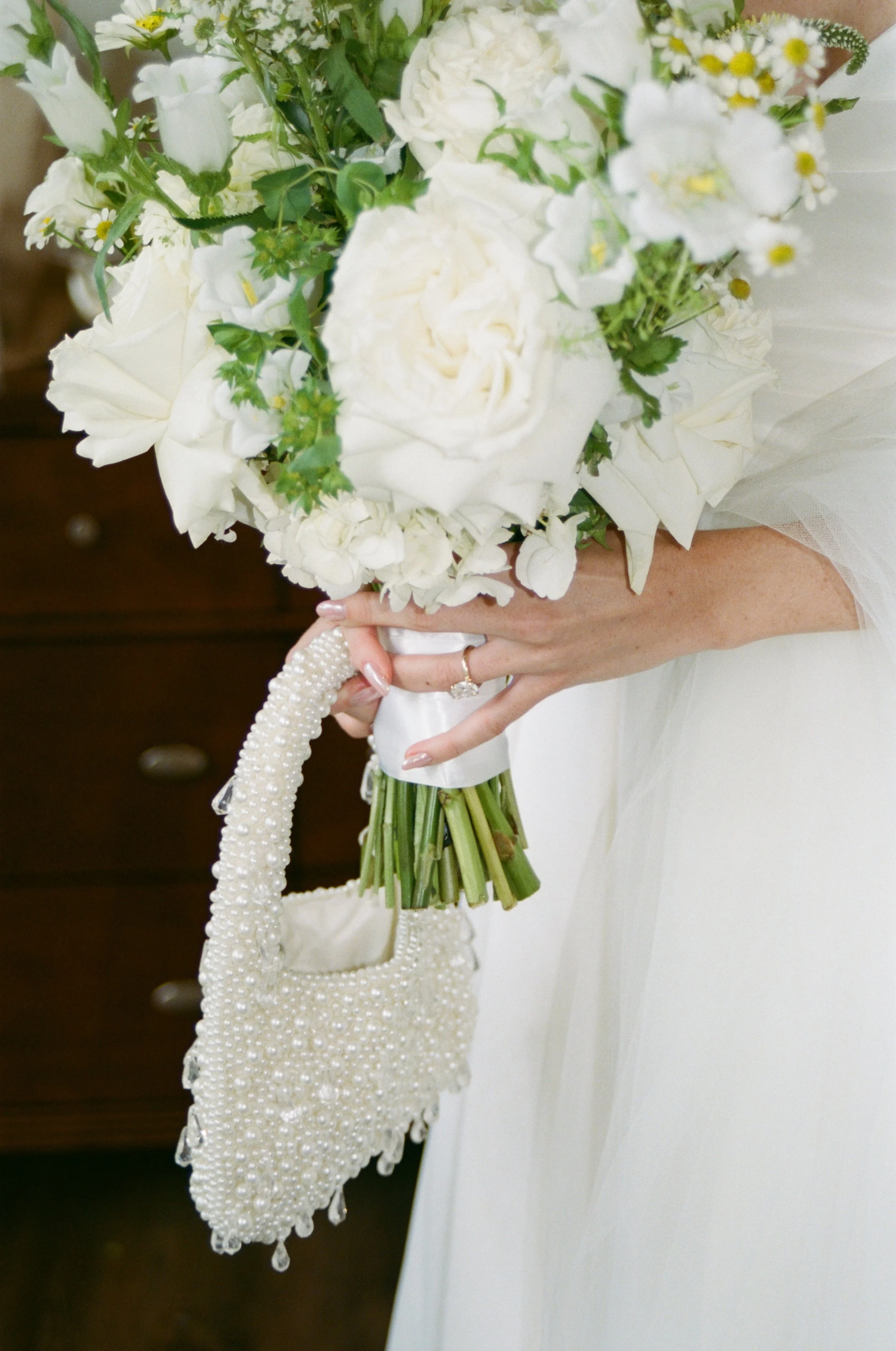 A bride holding a bouquet of white flowers, including roses and daisies, with a pearl-embellished handbag hanging from her hand, wearing a diamond ring and a white wedding dress.