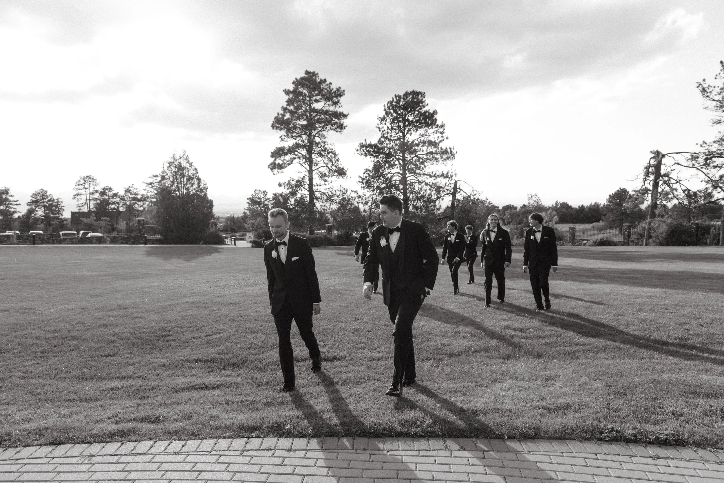 A group of groomsmen in tuxedos walking across a grassy field, with trees and a partly cloudy sky in the background.