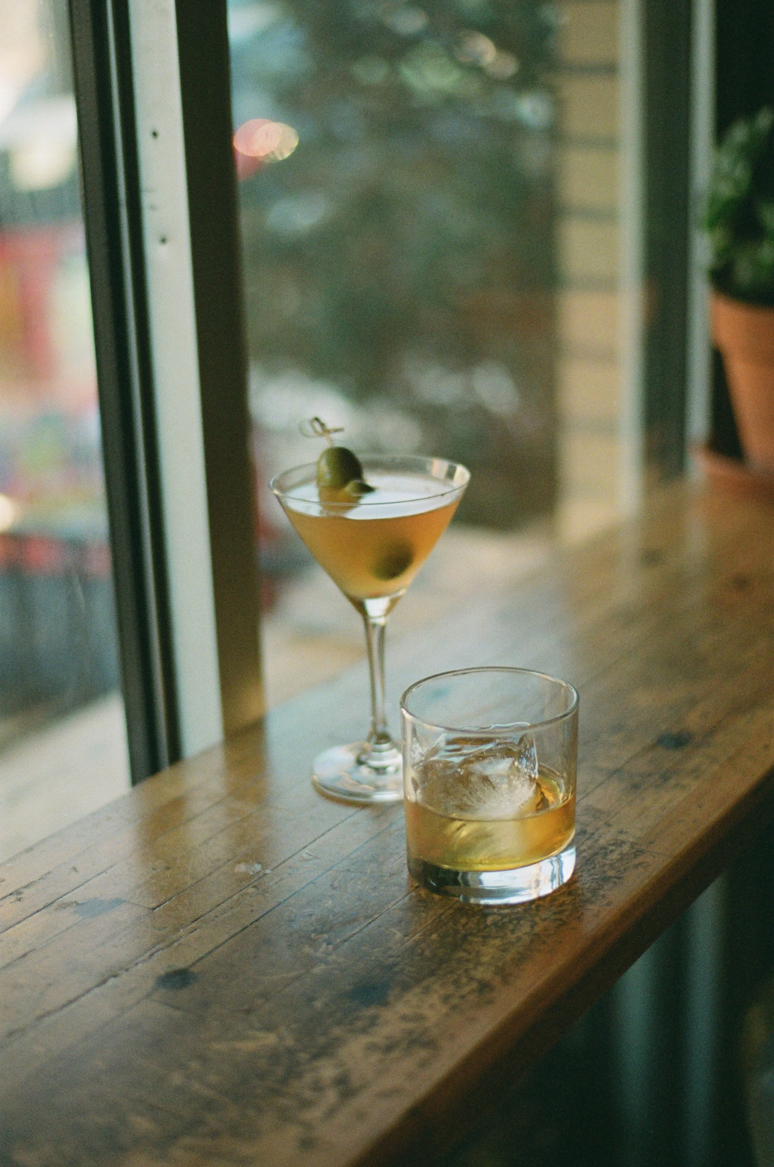 Two cocktails on a wooden bar counter, one martini glass with green olives and an old-fashioned glass with ice and a light-colored drink, near a large window.