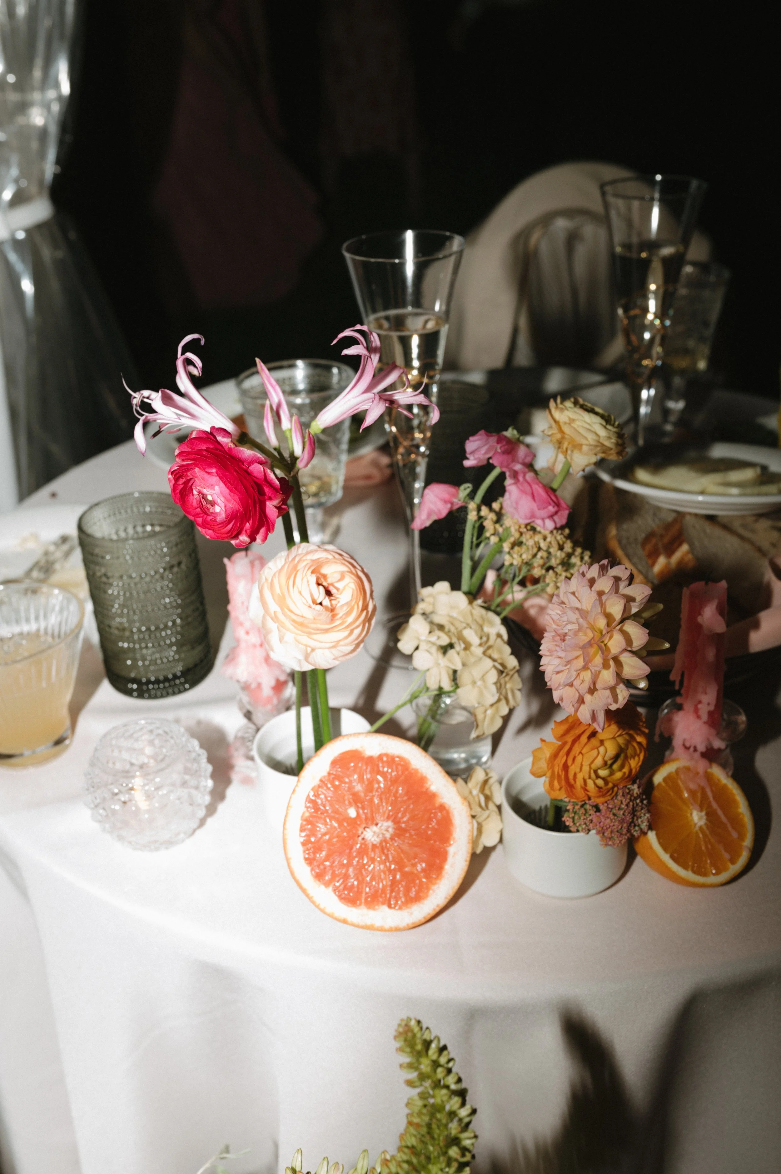 Table decorated with pink, peach, and yellow flowers in vases, sliced grapefruit and oranges, and glasses of champagne at a celebration or event.