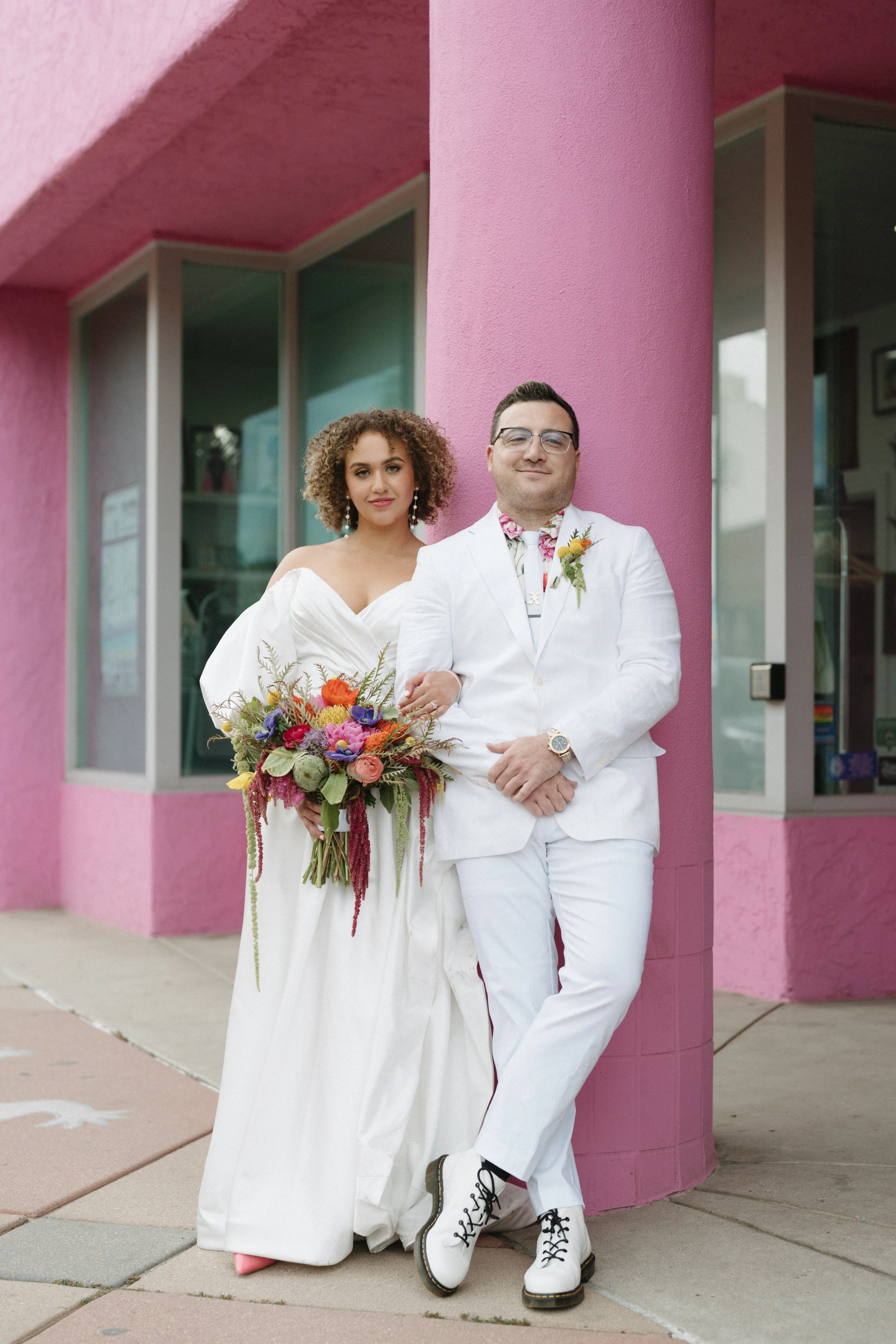 A bride and groom standing outside of a pink building on Santa Fe art district in Denver Colorado. bride has pink heels and the groom has white doc Martens. 