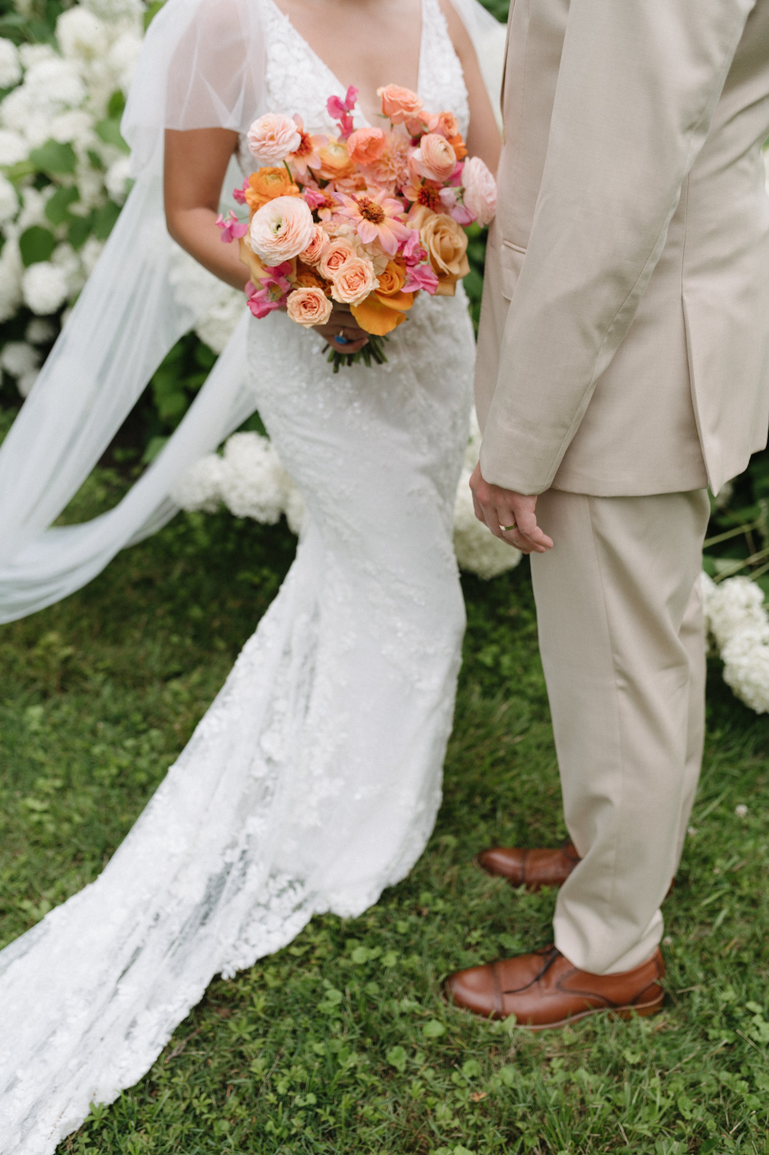Close-up of a bride and groom during their wedding ceremony, with the bride holding a colorful bouquet of pink and orange flowers, standing on green grass outdoors.