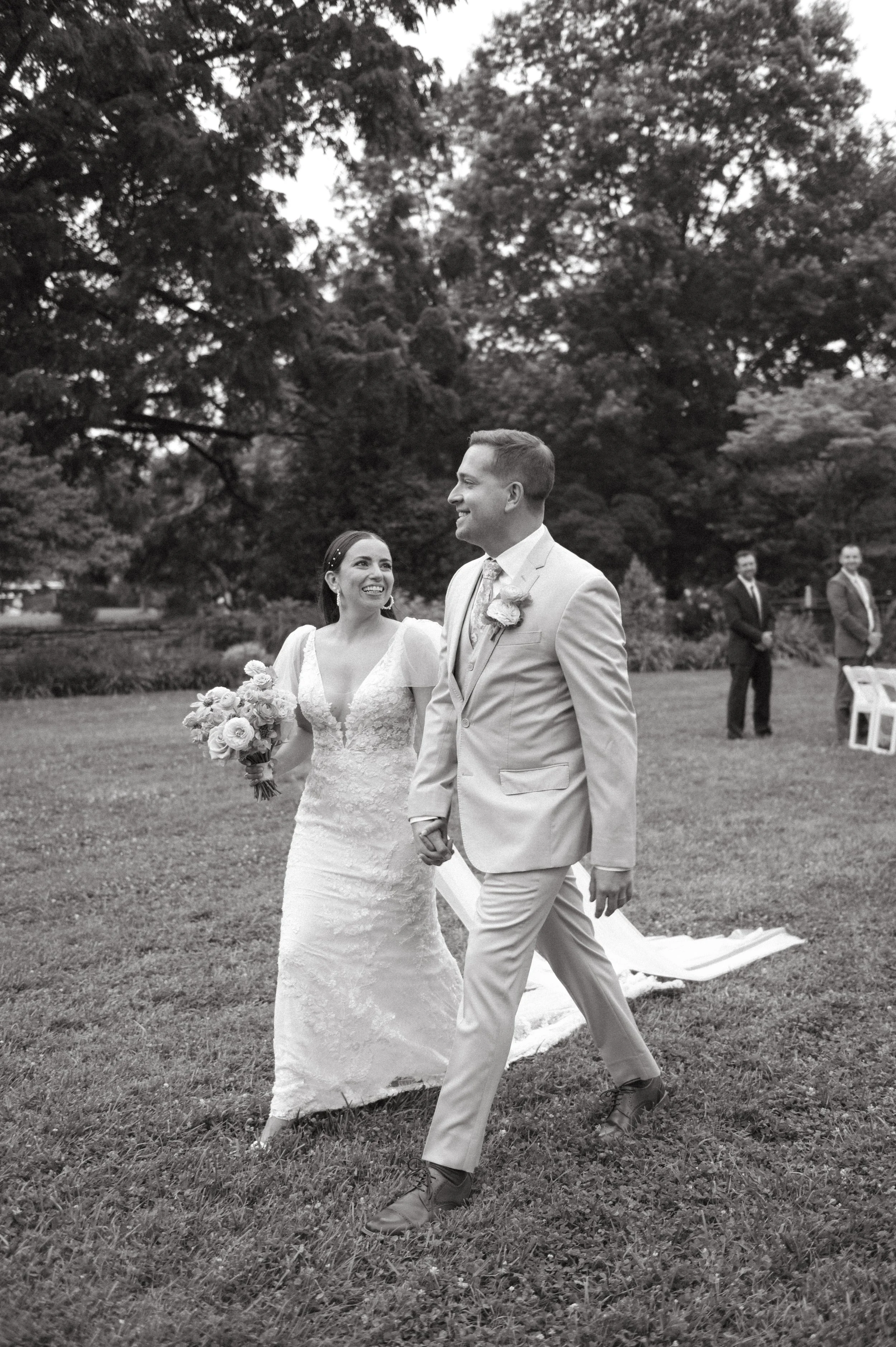 Black and white photo of a newlywed couple walking outdoors on a grassy area, holding hands and smiling at each other, with three men in suits standing in the background.