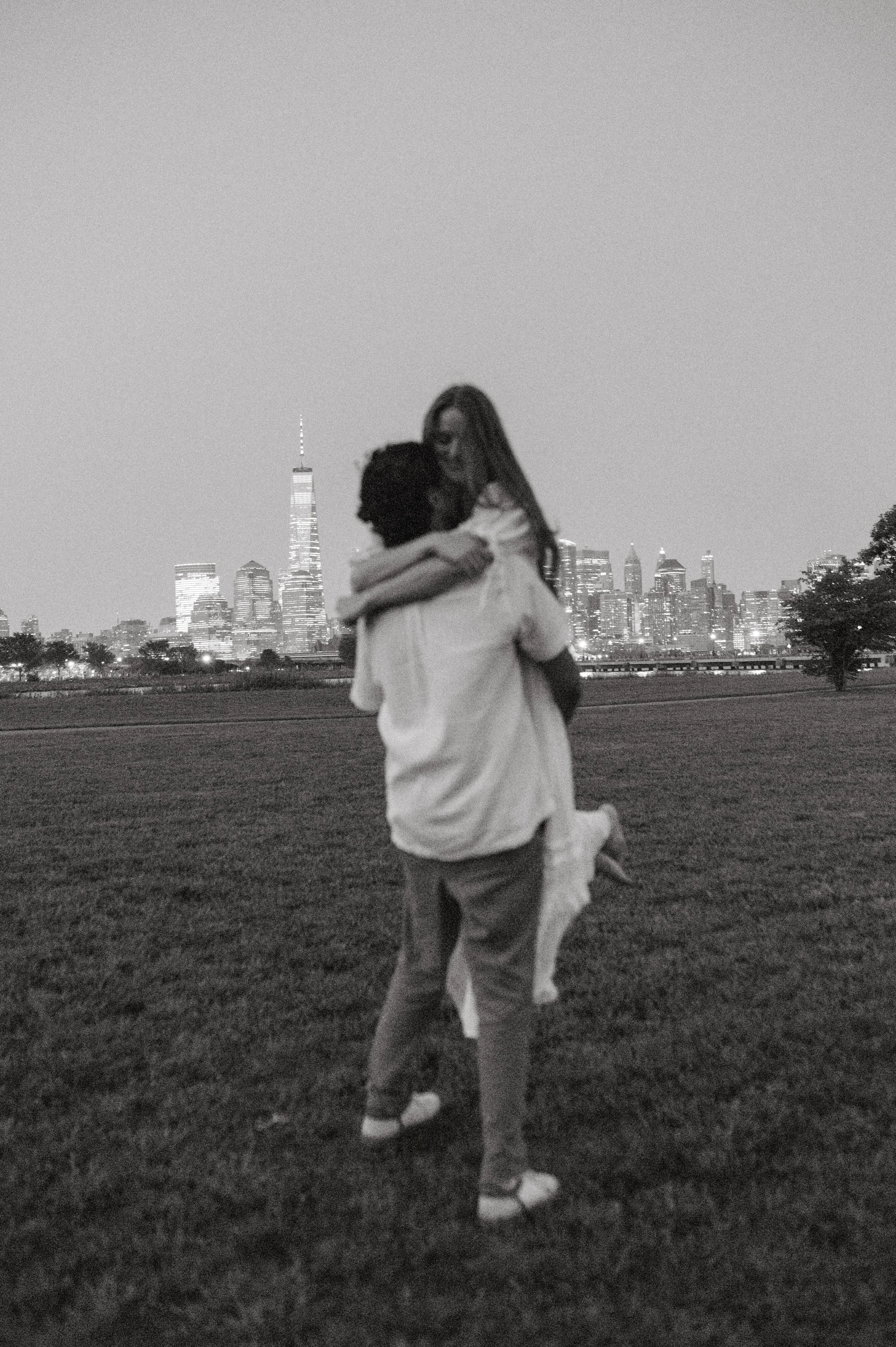A black and white photo of a couple embracing in a park at night with a city skyline, including the One World Trade Center, in the background.