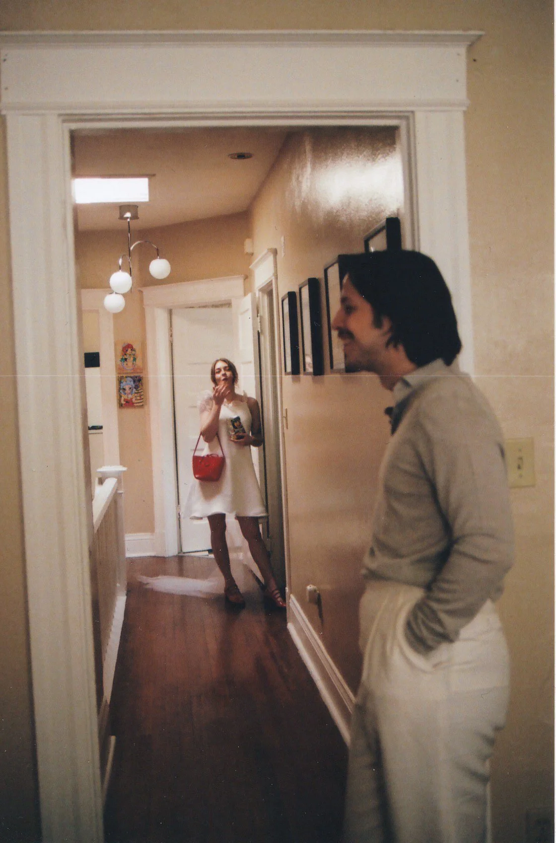 A groom in a custom suit standing with hands in pockets in a hallway, with his bride in a white dress and red purse smiling and blowing a kiss at him, seen through a doorway mirror.