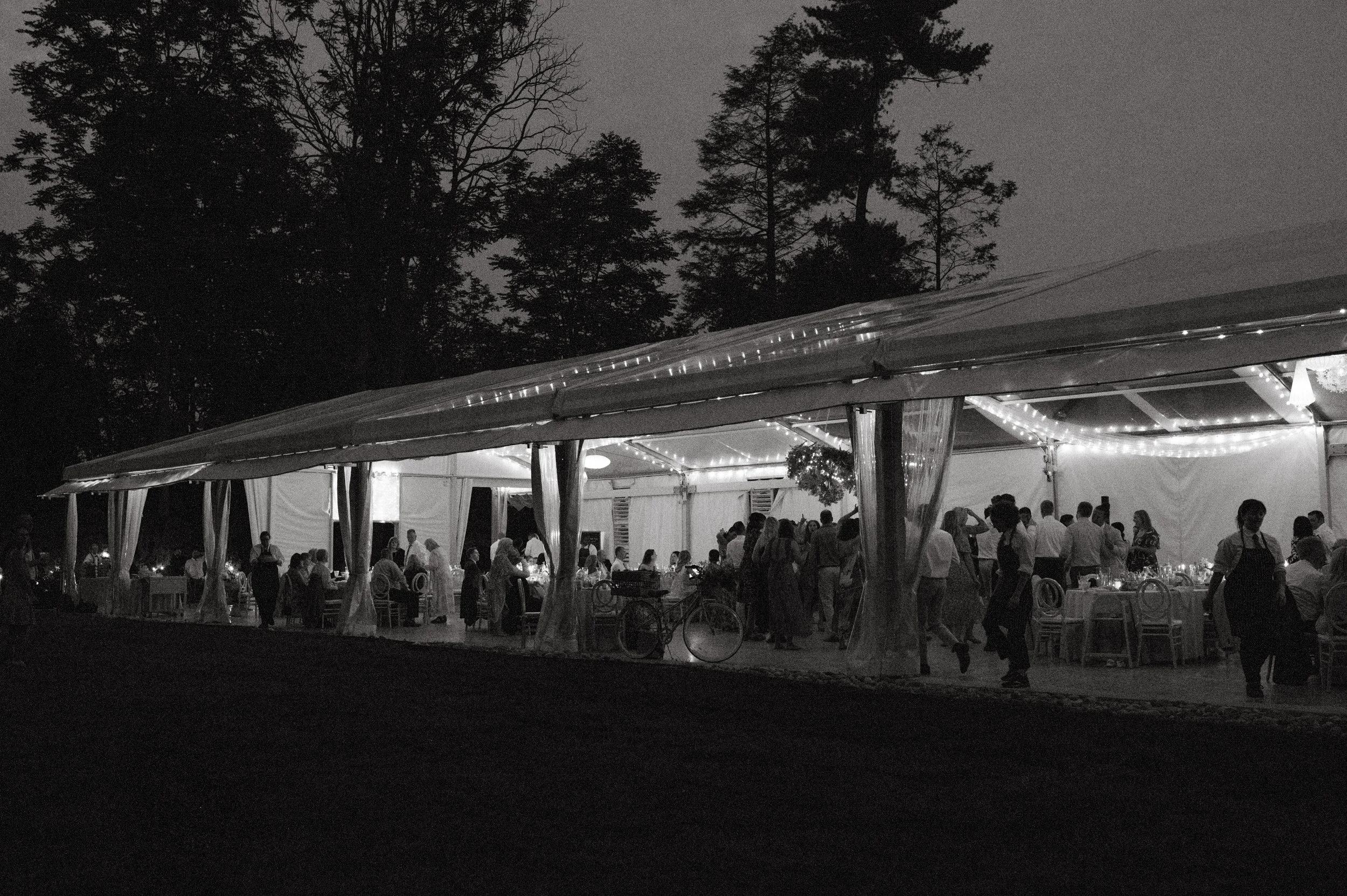 Nighttime outdoor reception with a large illuminated tent, people dining and socializing inside, surrounded by tall trees.