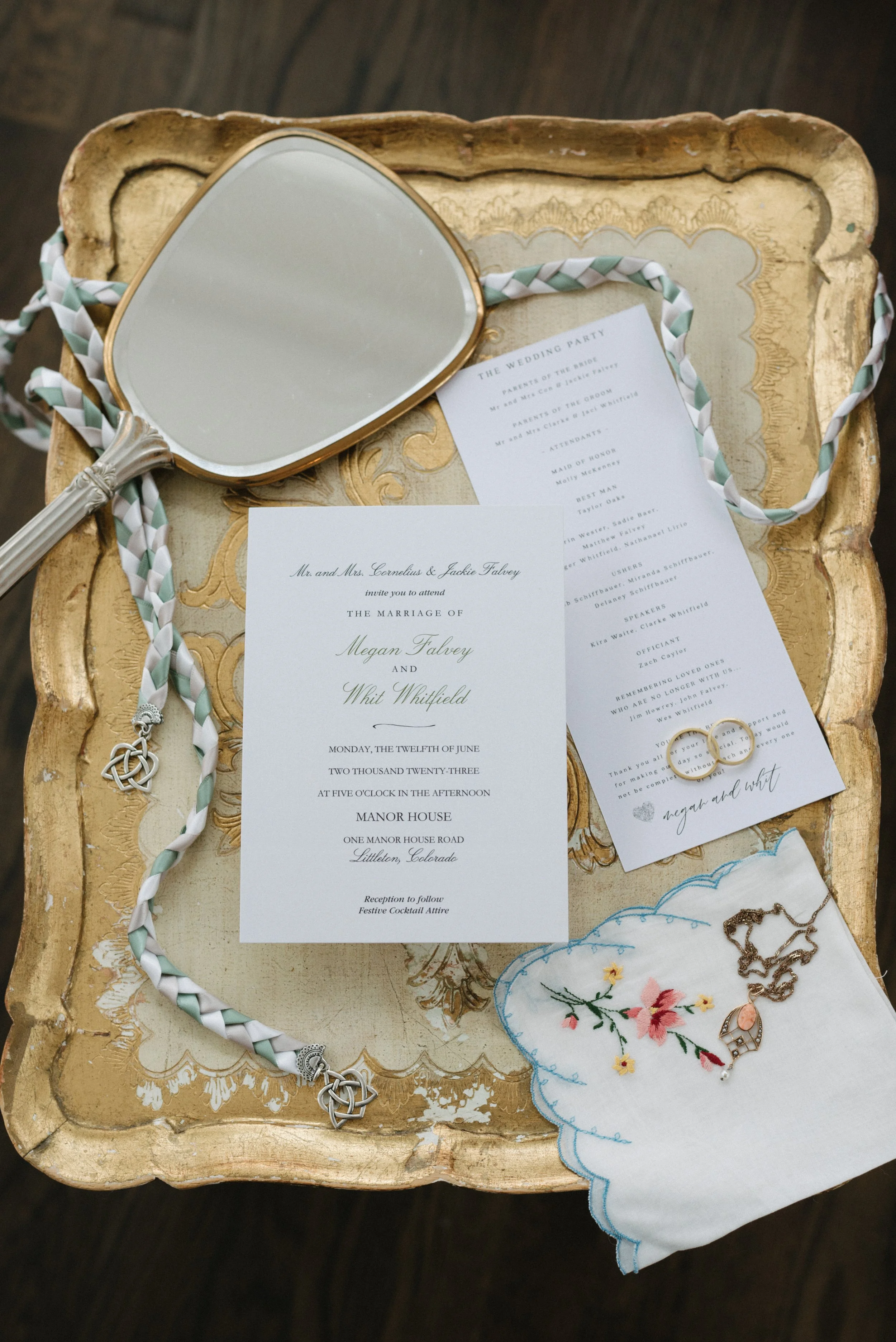 Wedding invitation suite on a vintage gold tray, with a hand mirror, embroidered handkerchief, jewelry, and a braided necklace.