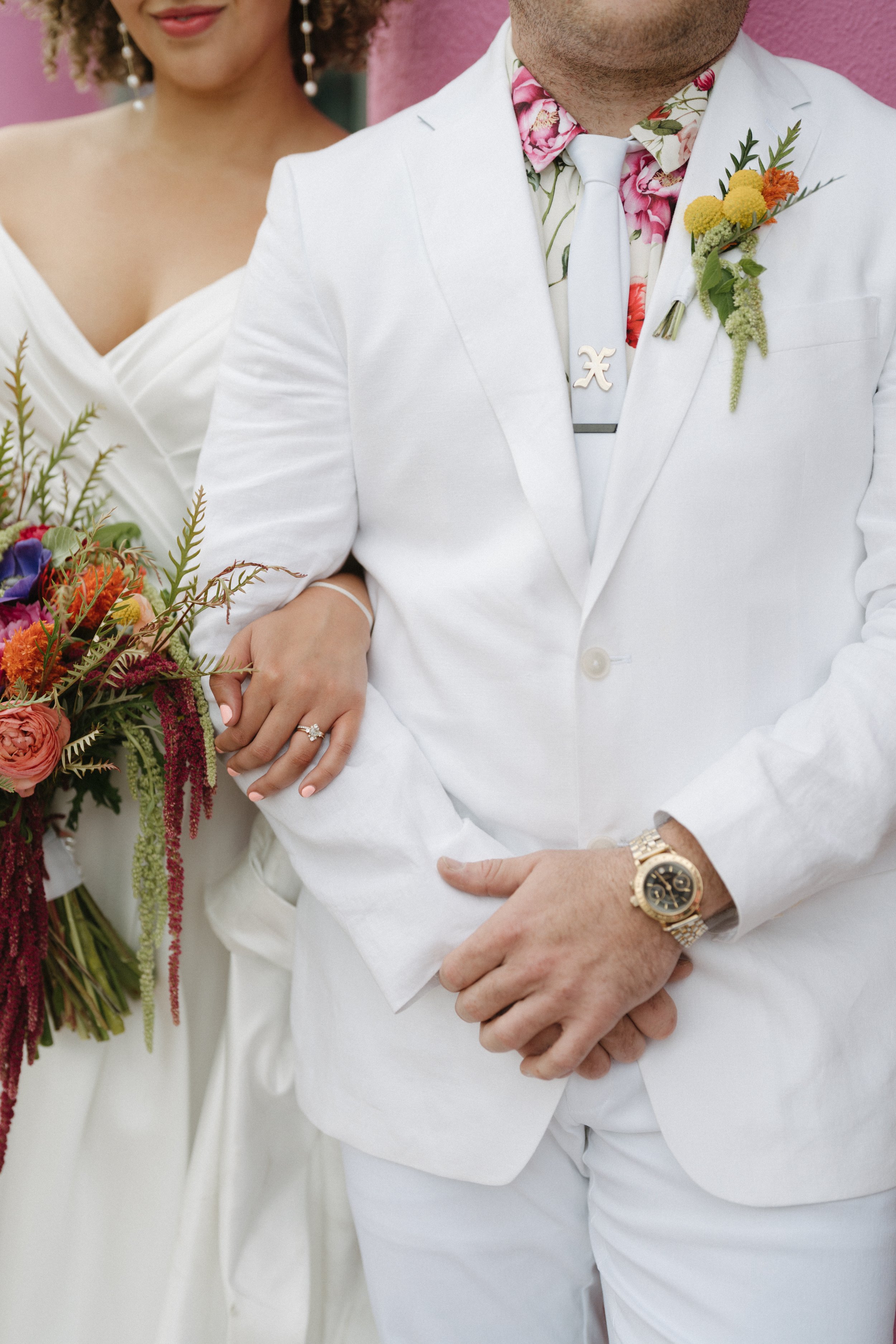 Close-up of a wedding couple, focusing on their torsos. The woman is wearing an off-the-shoulder white wedding dress, a ring on her left hand, and is holding a bouquet of colorful flowers. The man is dressed in a white suit with a floral shirt undern