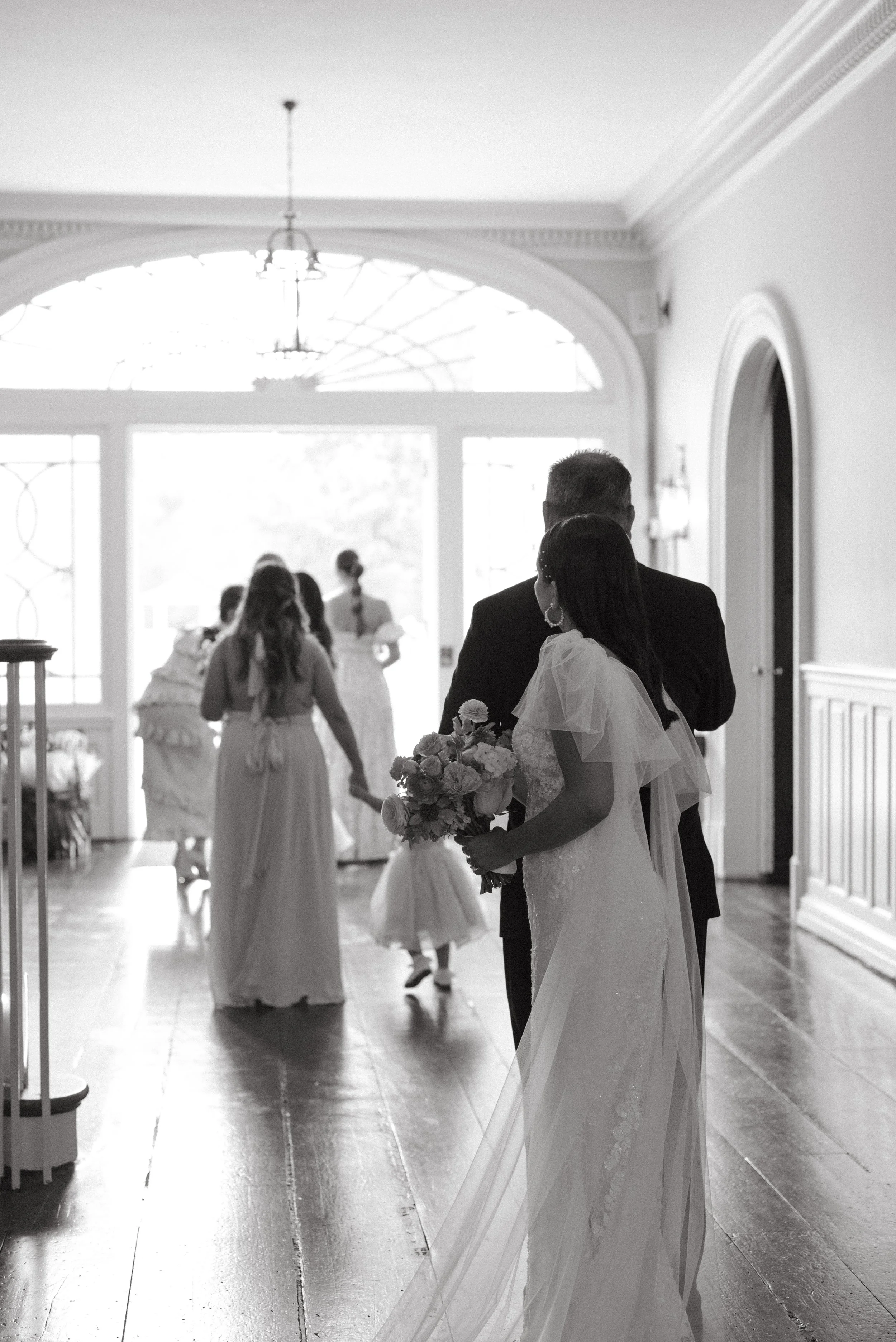Black and white photo of a bride holding a bouquet walking into a wedding venue, with guests and a woman in the background near the door.