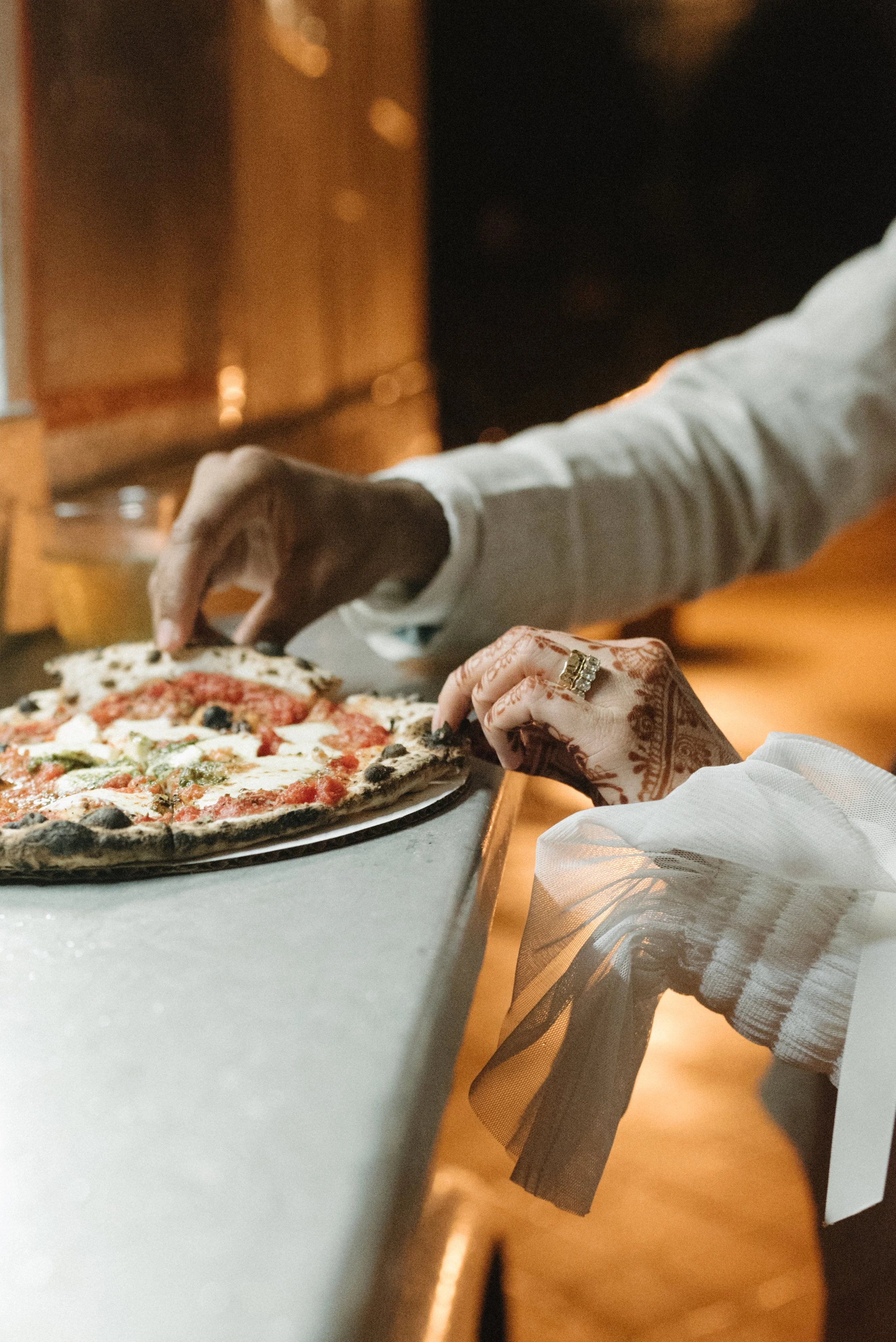 Bride and Grooms hands reaching for a slice of pizza at a food truck during their reception. The bride has  henna tattoos on the right hand and a wedding ring.