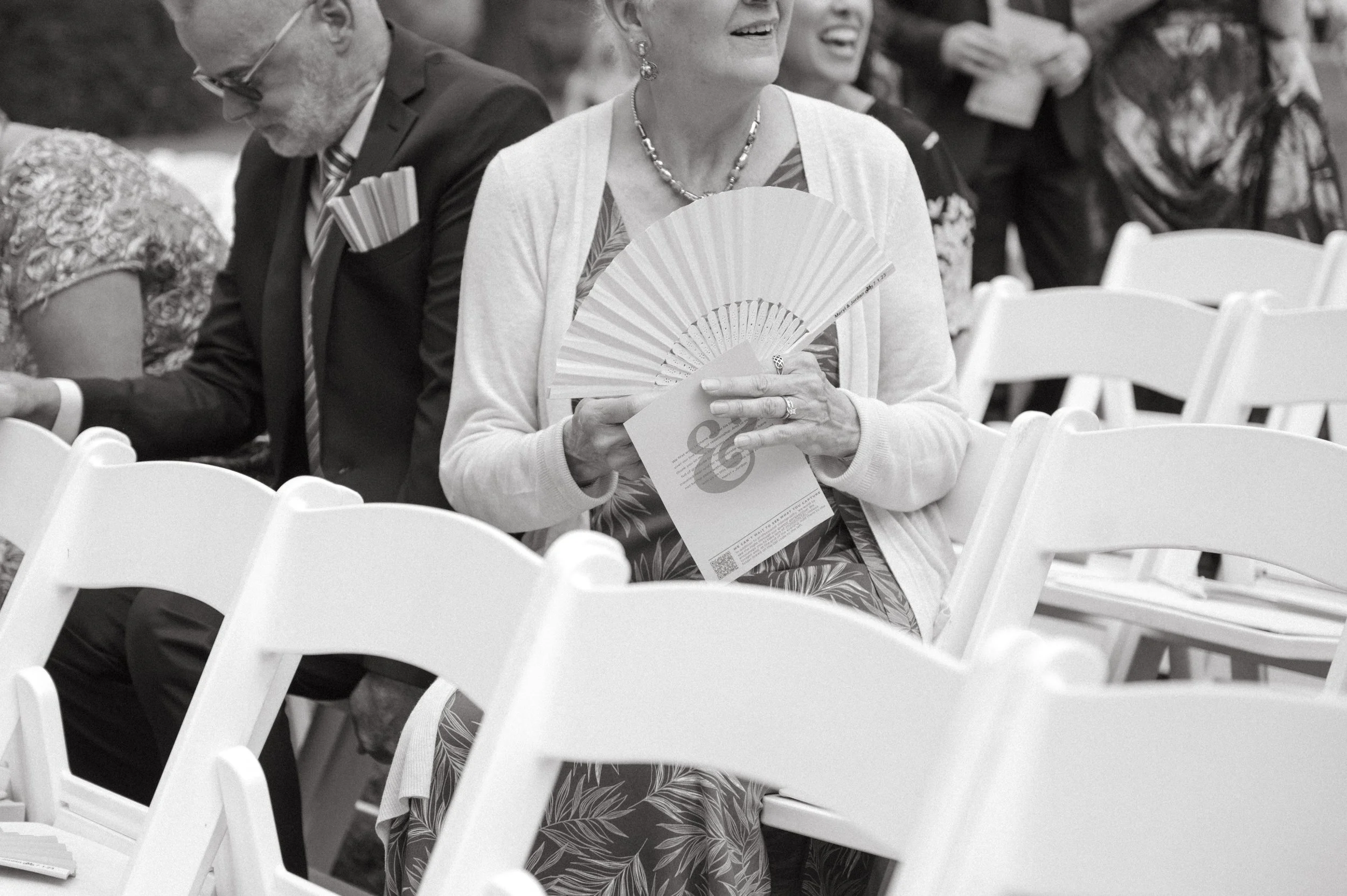  the brides grandmother seated holding a costume fan watching the bride walk down the isle at the summer outdoor wedding. 