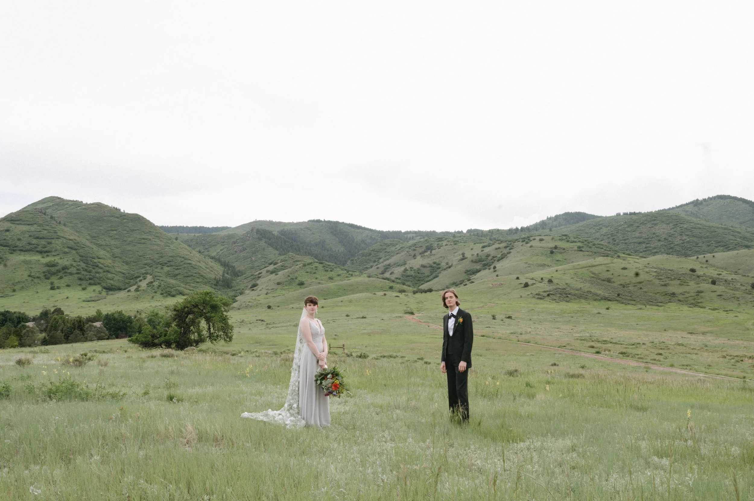 A bride and groom standing apart in a green field with hills and trees in the background, during a wedding photo shoot.