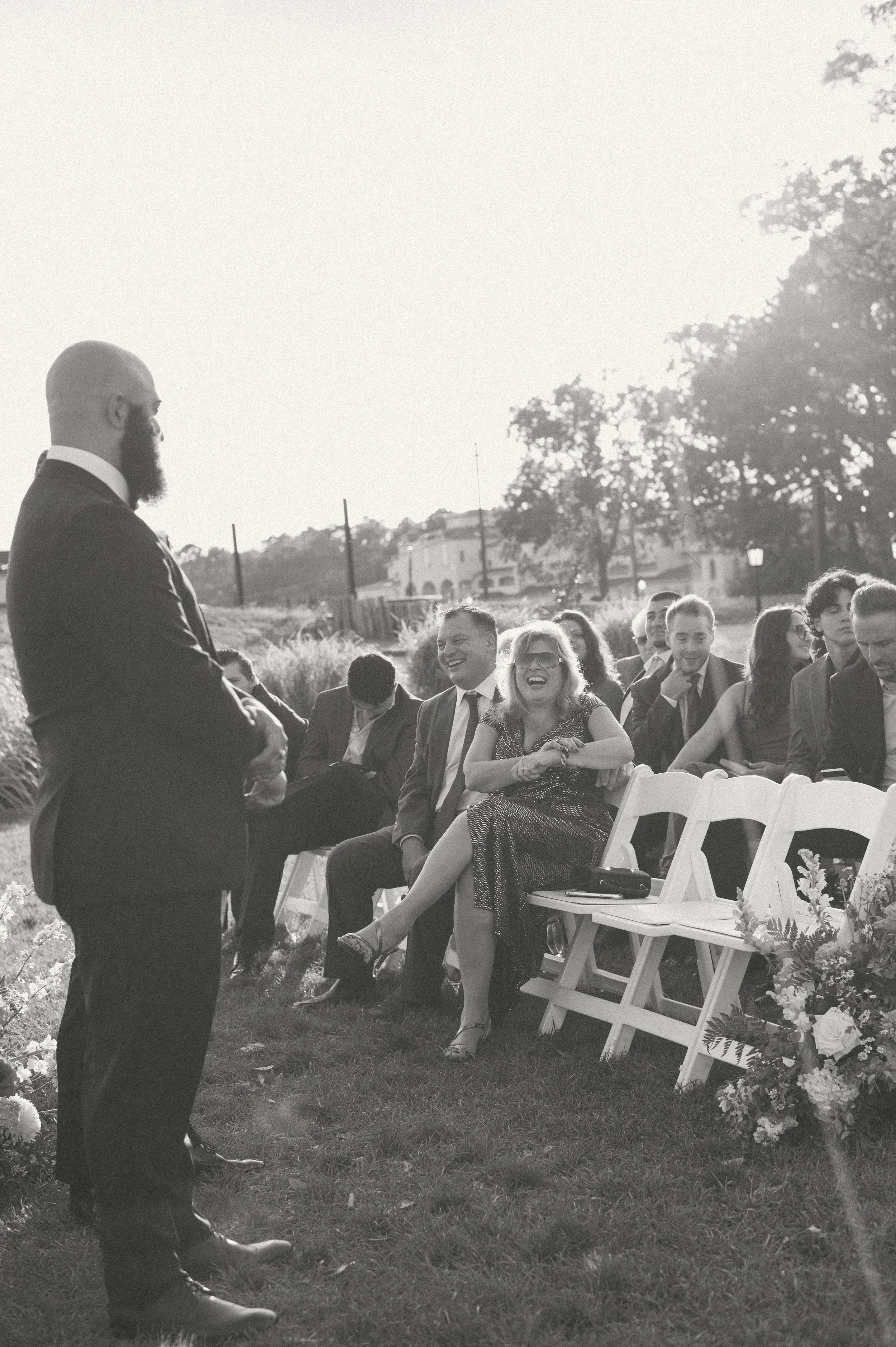A wedding ceremony outdoors with guests seated on white chairs, some laughing and smiling, as a man in a suit stands speaking.