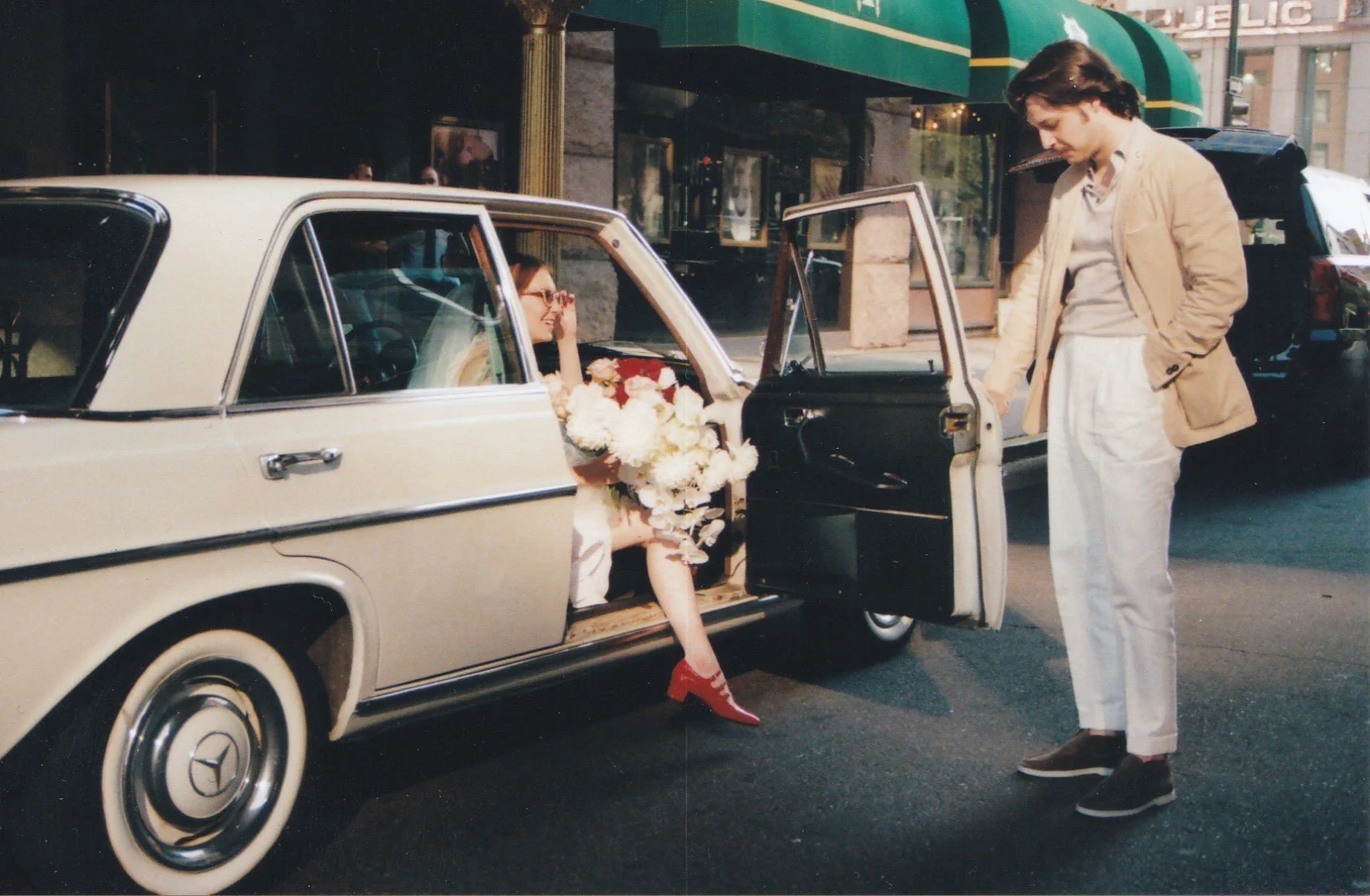 A Bride holding a bouquet of white flowers sitting in a vintage beige Mercedes-Benz car, while her groom in white pants and a beige jacket stands outside, looking into the car, on a city street in front of the brown palace downtown Denver.