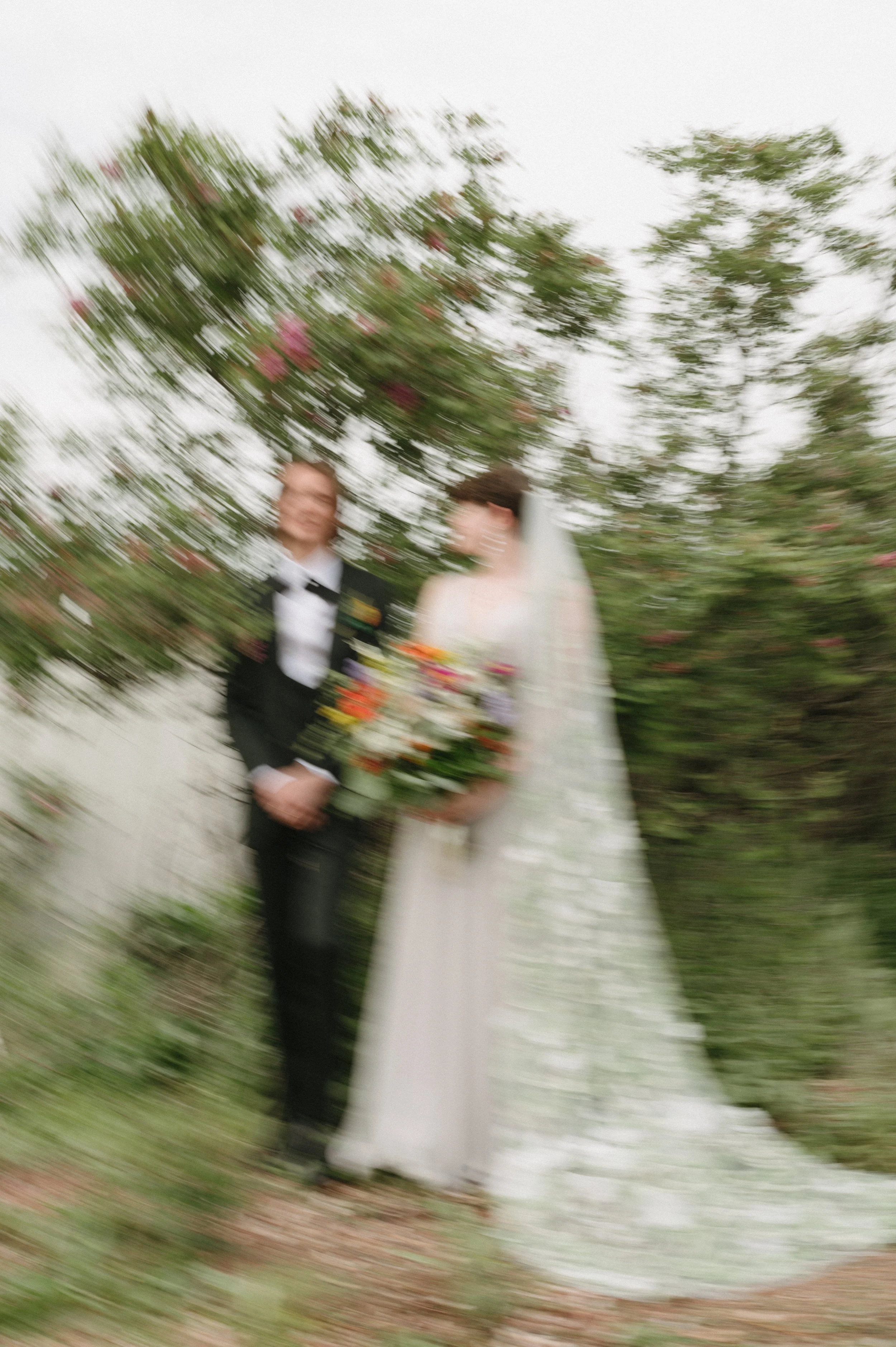 Blurred photo of a bride in a white wedding dress and veil holding a bouquet, standing next to a groom in a tuxedo, outdoors with trees and pink flowers in the background.