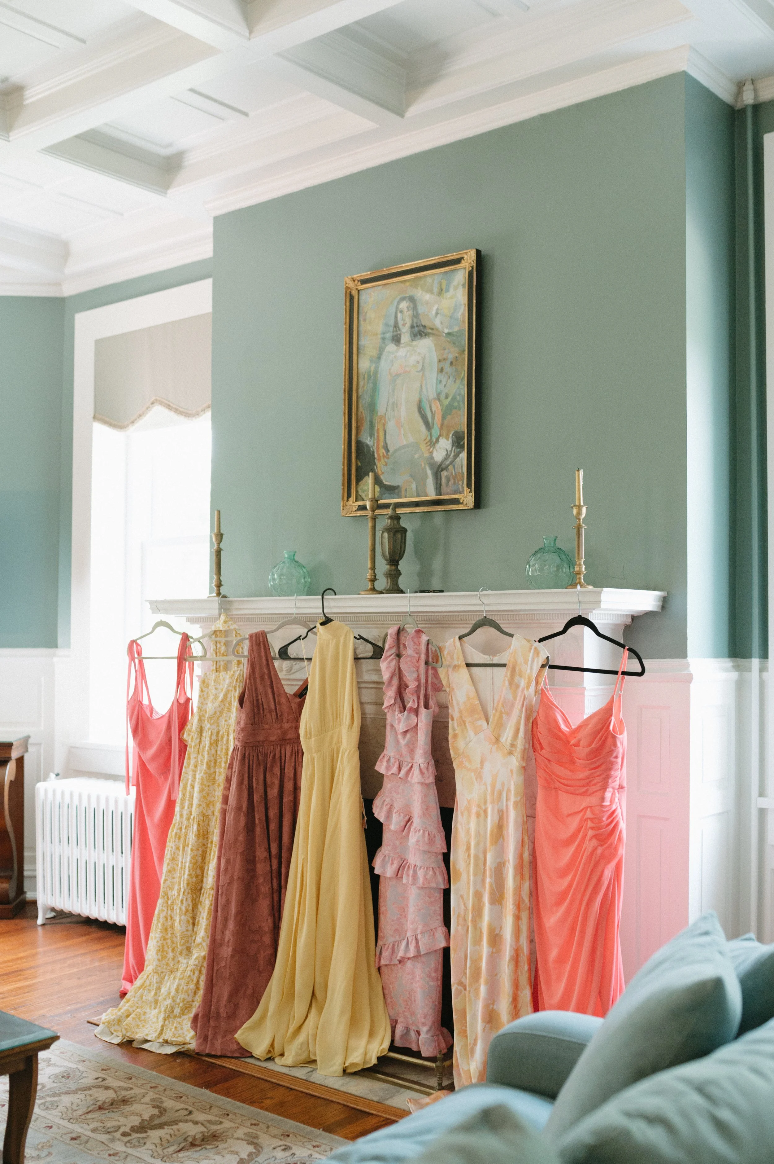 bridesmaids colorful dresses hanging on a metal rack in front of a fireplace with a green wall, topped with candles and decorative glass vases, and a framed painting above.