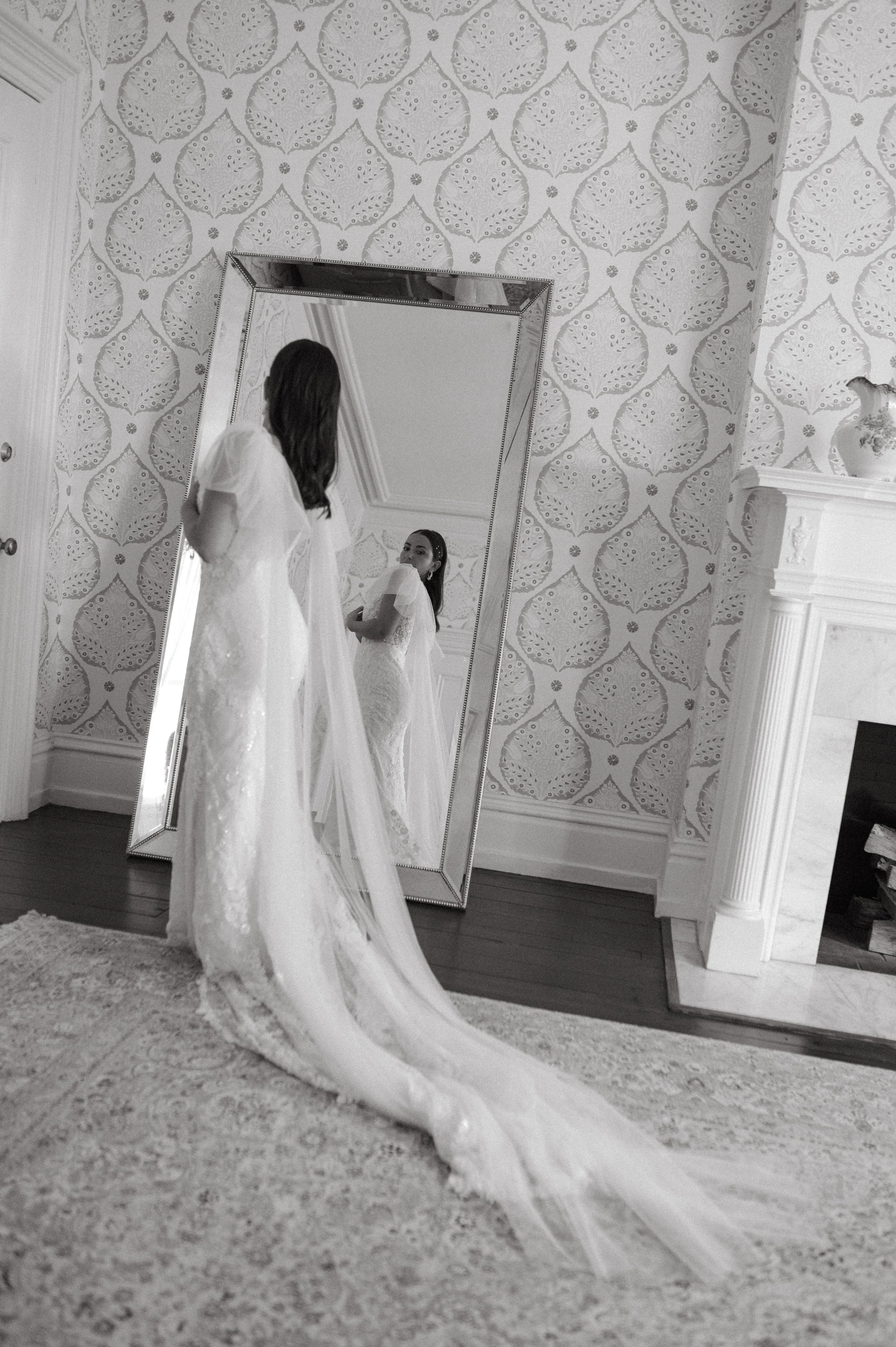 A woman in a wedding dress standing in front of a large mirror in a decorated room with a patterned wallpaper and a fireplace.