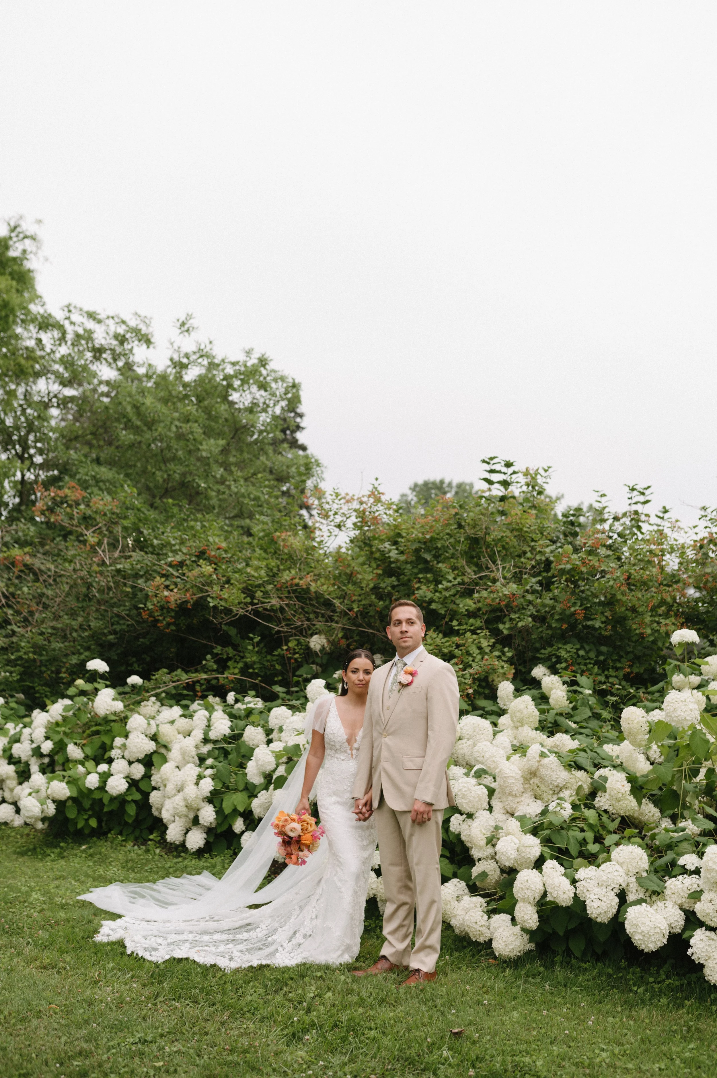 A bride and groom holding hands outdoors among white flowering bushes, dressed in wedding attire.