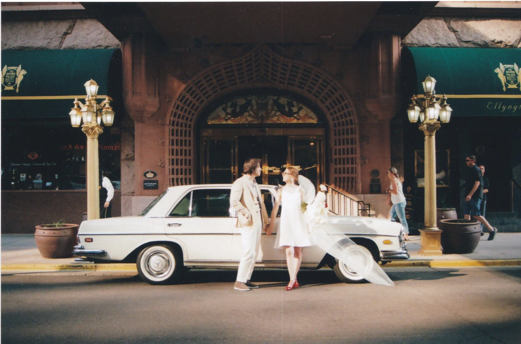 Bride and Groom holding hands in front of a white vintage car parked on the street, with a building entrance and pedestrians in the background. this was in front of the brown palace. 