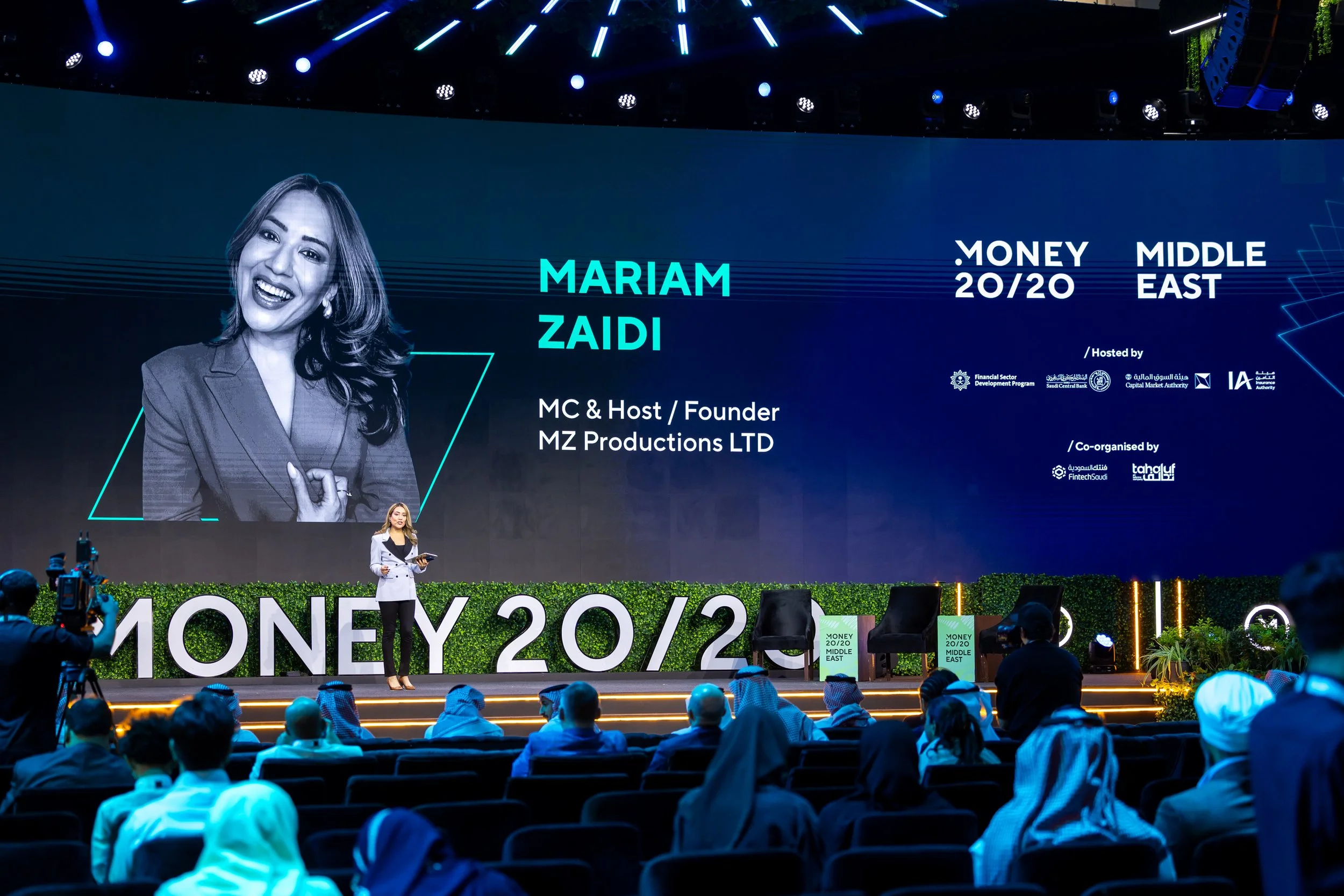 A woman standing on stage during a conference, with a large screen behind her displaying her name, Mariam Zaidi, and details about the event, Money 20/20 Middle East. She is holding a tablet and speaking to an audience.