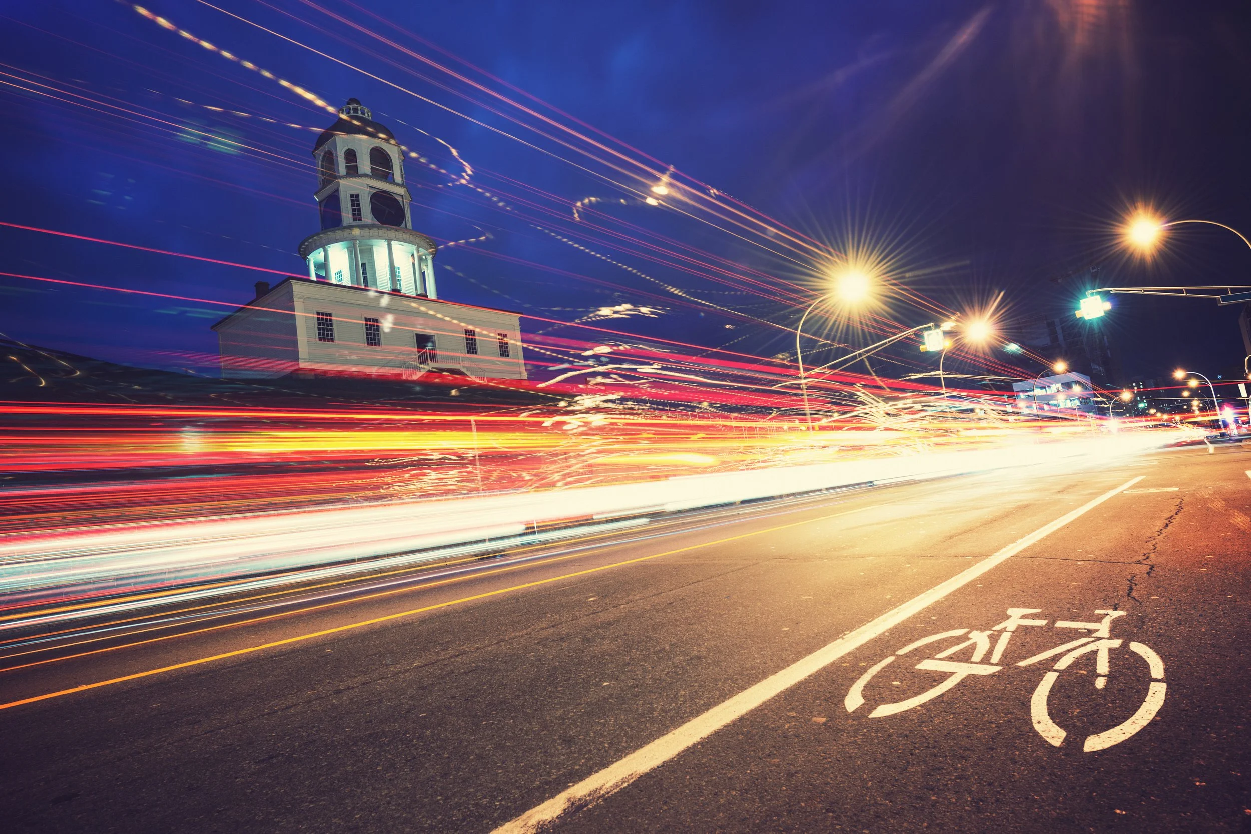 Nighttime city scene with a historic clock tower, light trails from passing vehicles on the street, and a designated bicycle lane with a bicycle symbol painted on the pavement.