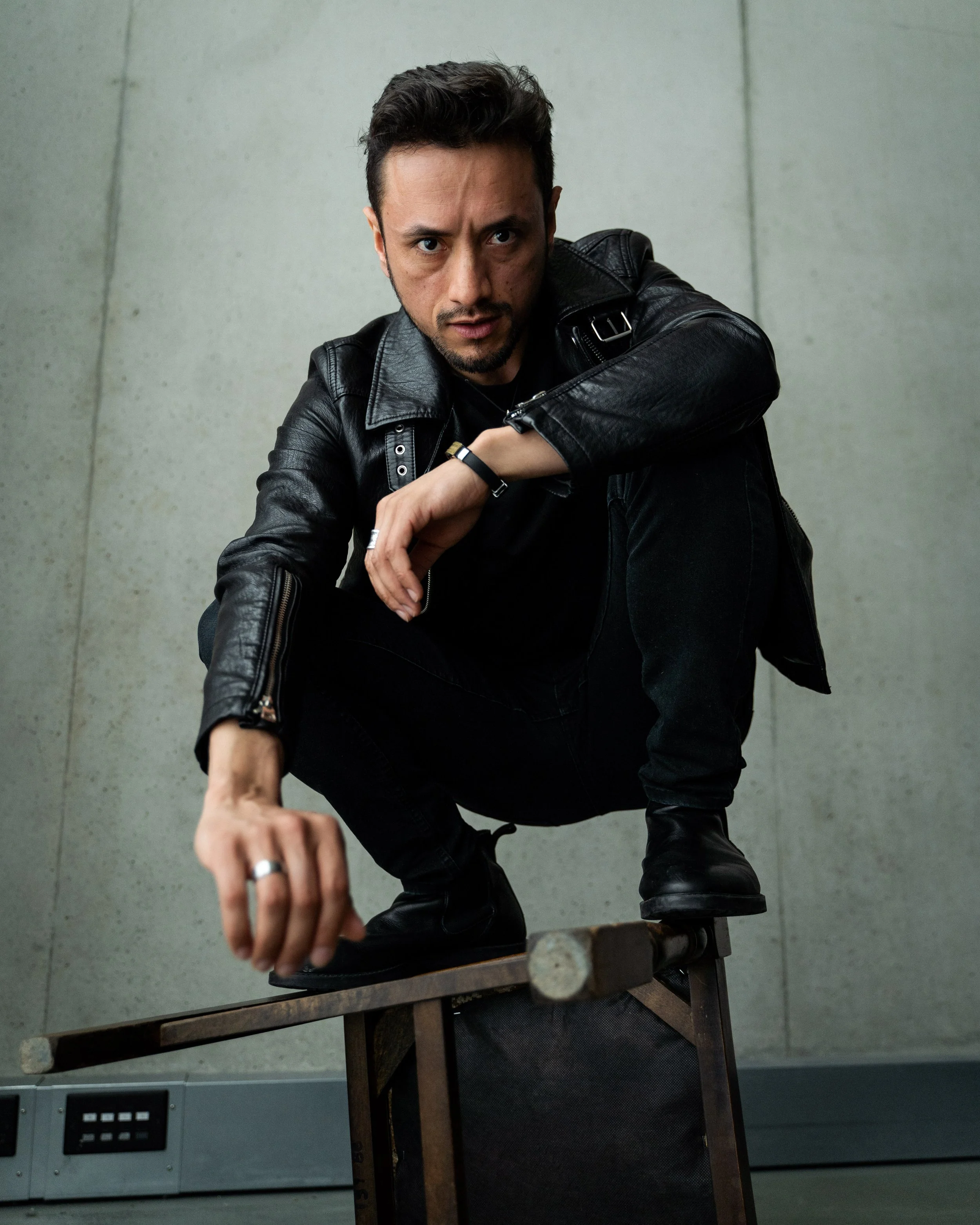 Miguel Torres Umba crouching on a chair with one hand on the floor, wearing a black leather jacket and black pants, looking directly at the camera.