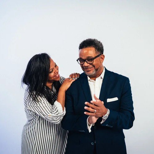 A woman and a man are smiling and standing close together. The woman, wearing a striped shirt, has her hand on the man's shoulder and appears to be playful and happy, while the man, in a dark suit and glasses, is smiling and holding his hands together.