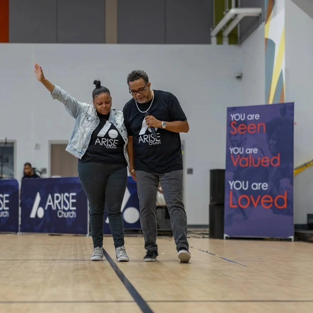 Two people, a man and a woman, standing on a stage or open area with a wooden floor, participating in a church event for Arise Church. The woman has her eyes closed, with one arm raised, and the man is holding a microphone while looking down at the floor. Both are wearing black T-shirts with the Arise Church logo. There are banners in the background with motivational messages: 'You are Seen, Valued, Loved,' and another banner displaying the Arise Church logo.