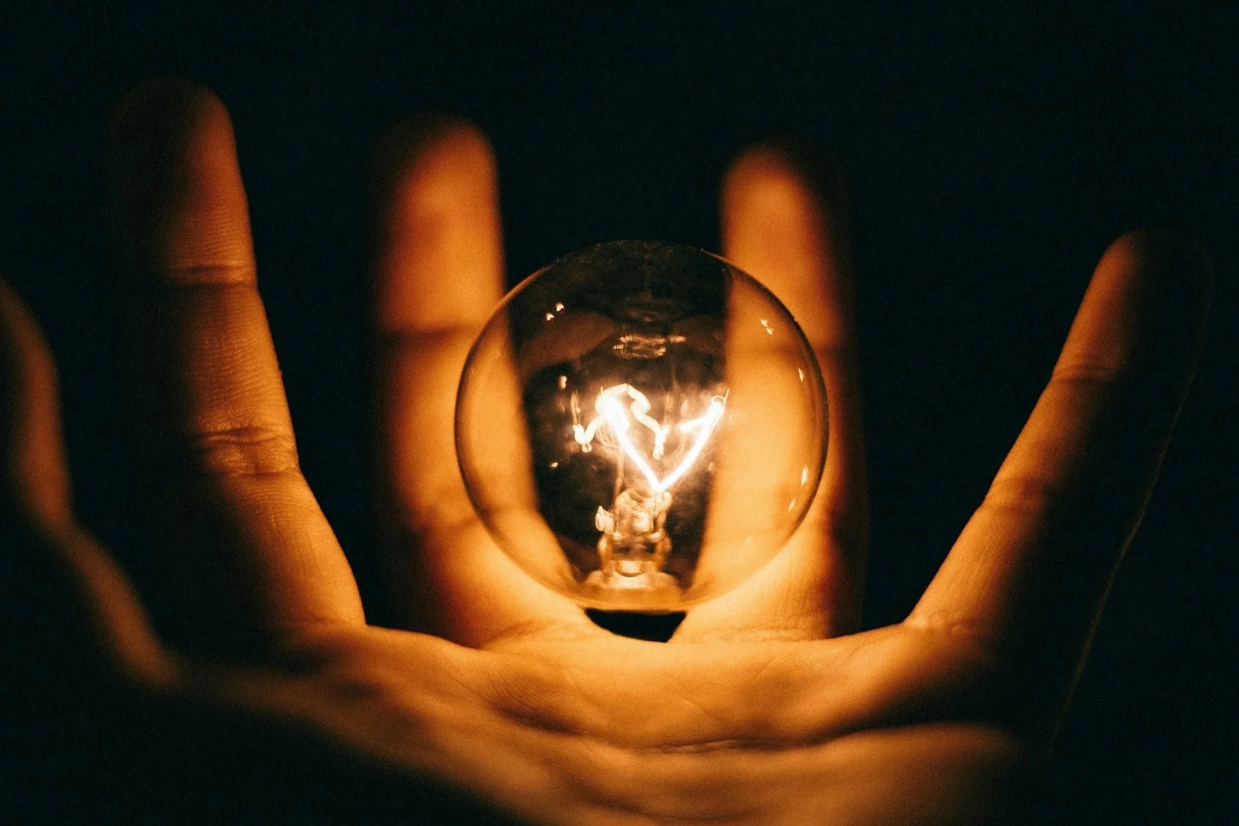 A hand holding a glass orb with a glowing light bulb inside, illuminating the dark background.