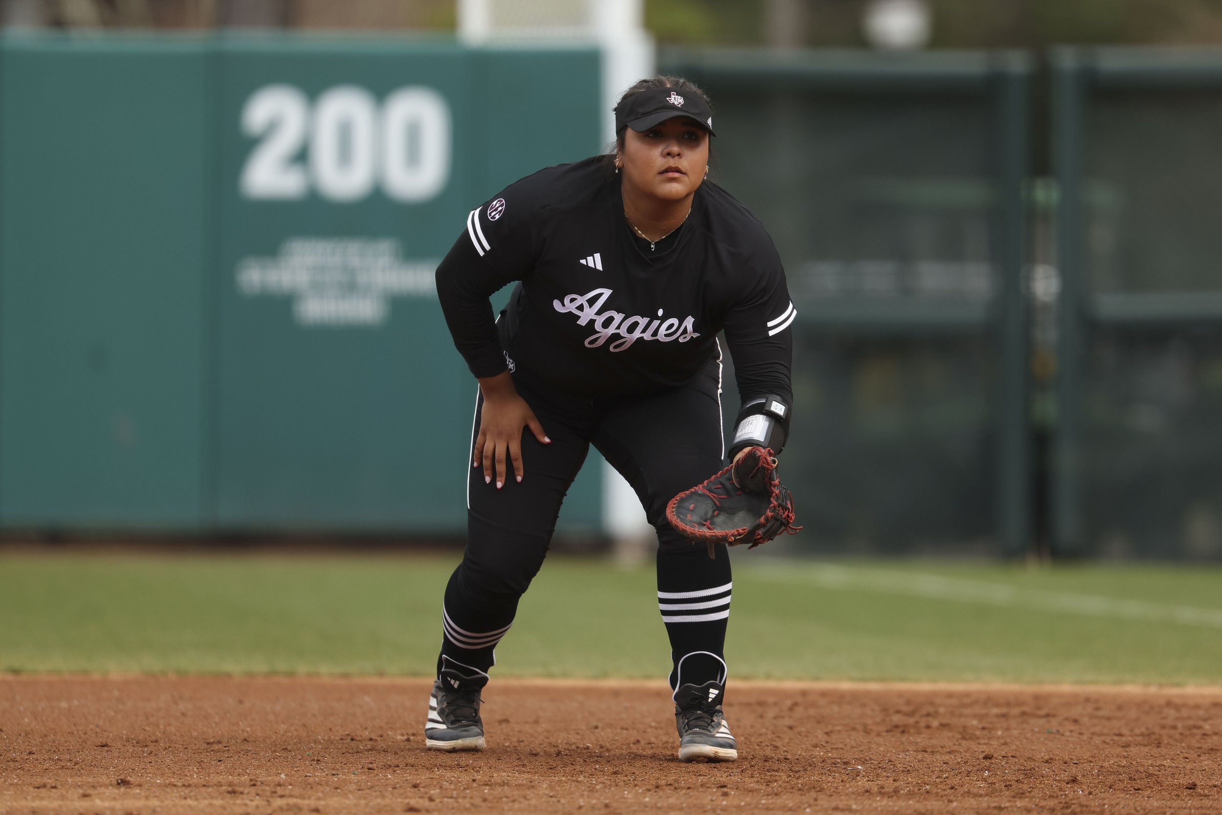 Mya Perez, Aggie Softball player, is crouched in a ready position on the field, wearing a black uniform with 'Aggies' written on it, a black cap, and holding a glove.