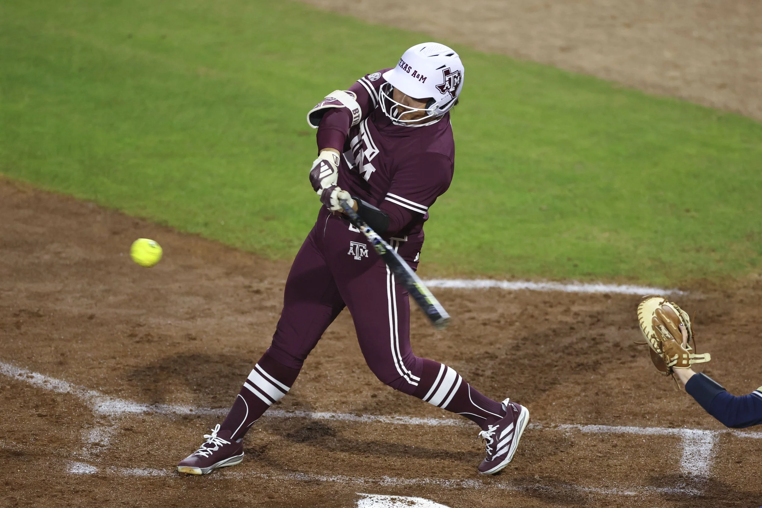 Mya Perez, Aggie Softball player, in a maroon uniform is swinging a bat at a yellow ball on the dirt infield of a softball field.
