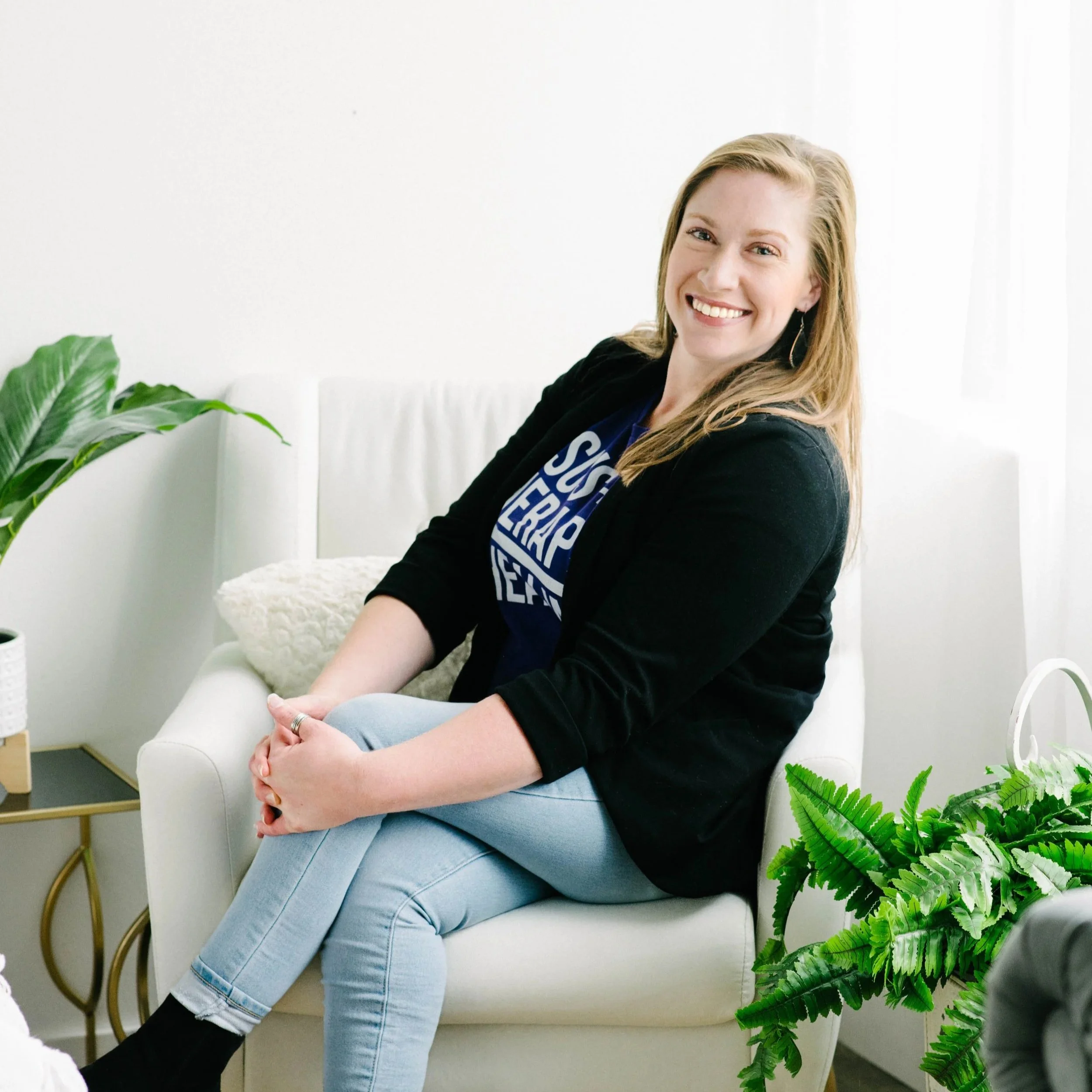 A woman with long brown hair smiling and sitting on a white armchair in a bright, modern room with green plants.