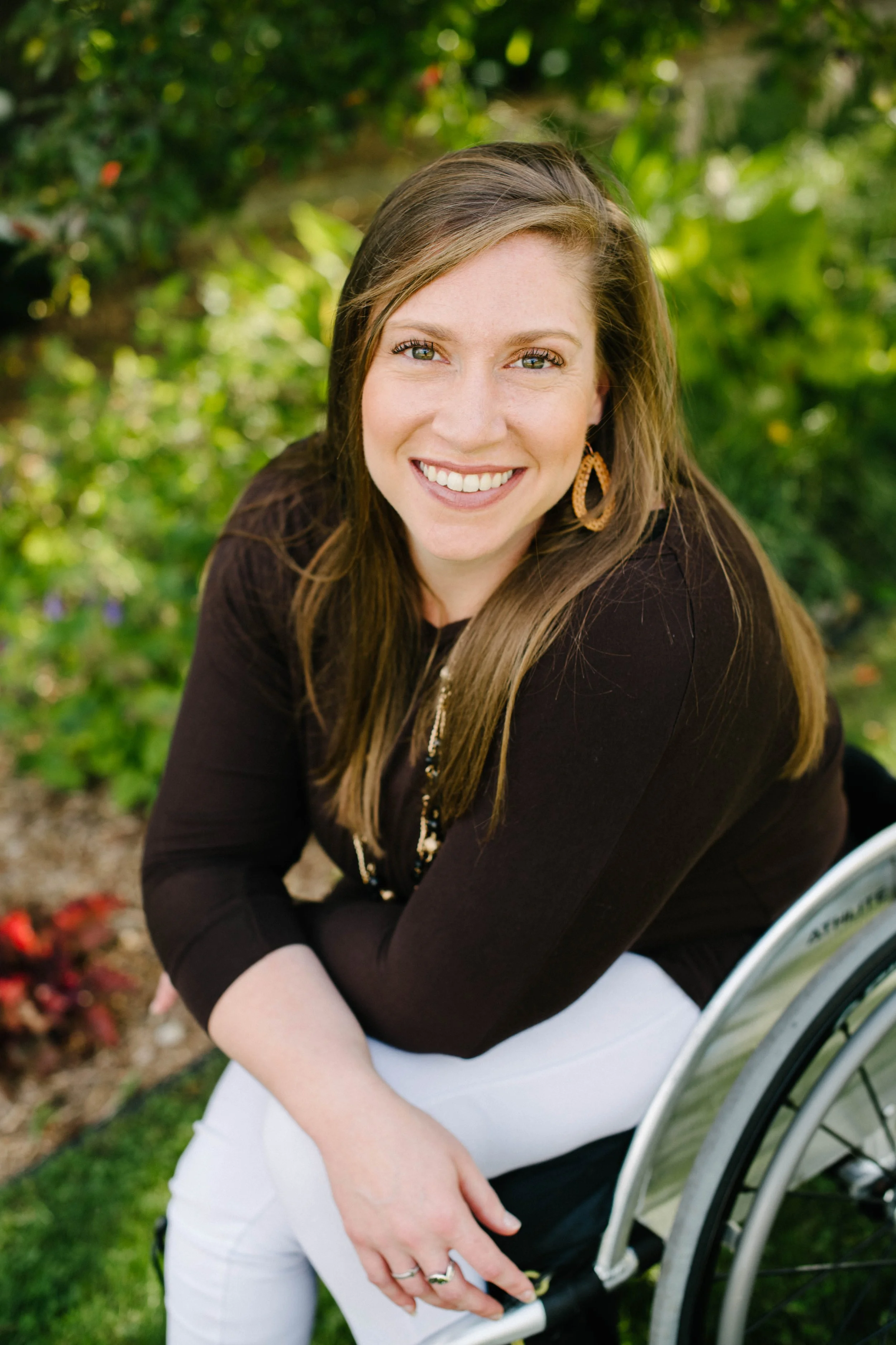 A smiling woman with long brown hair and blue eyes, wearing a dark long-sleeve top, white pants, and gold earrings, sitting in a wheelchair outdoors with green foliage in the background.