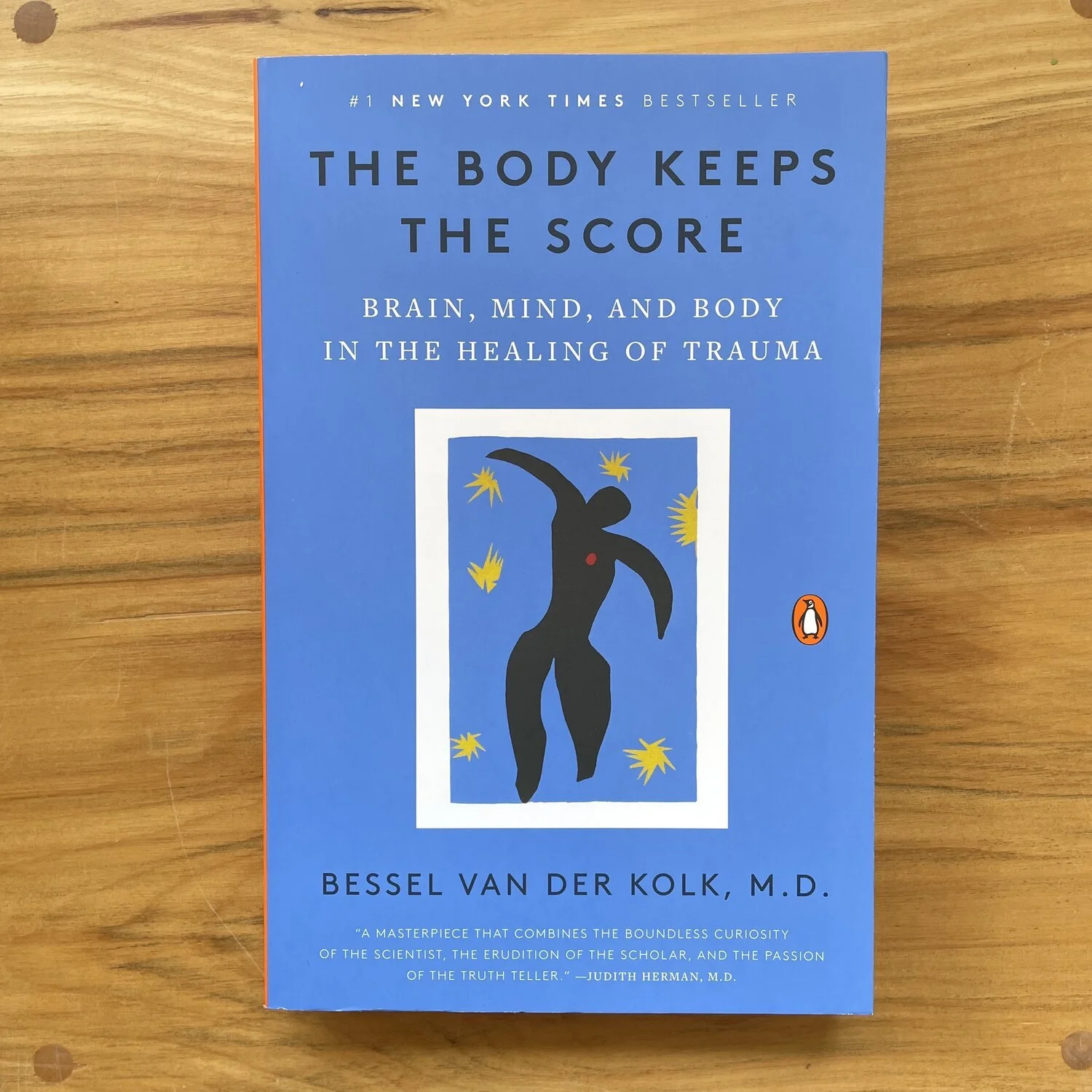 Book titled 'The Body Keeps the Score' by Bessel Van Der Kolk, placed on a wooden surface. The cover is blue with an abstract black figure and yellow star-like shapes, and features the Penguin logo.