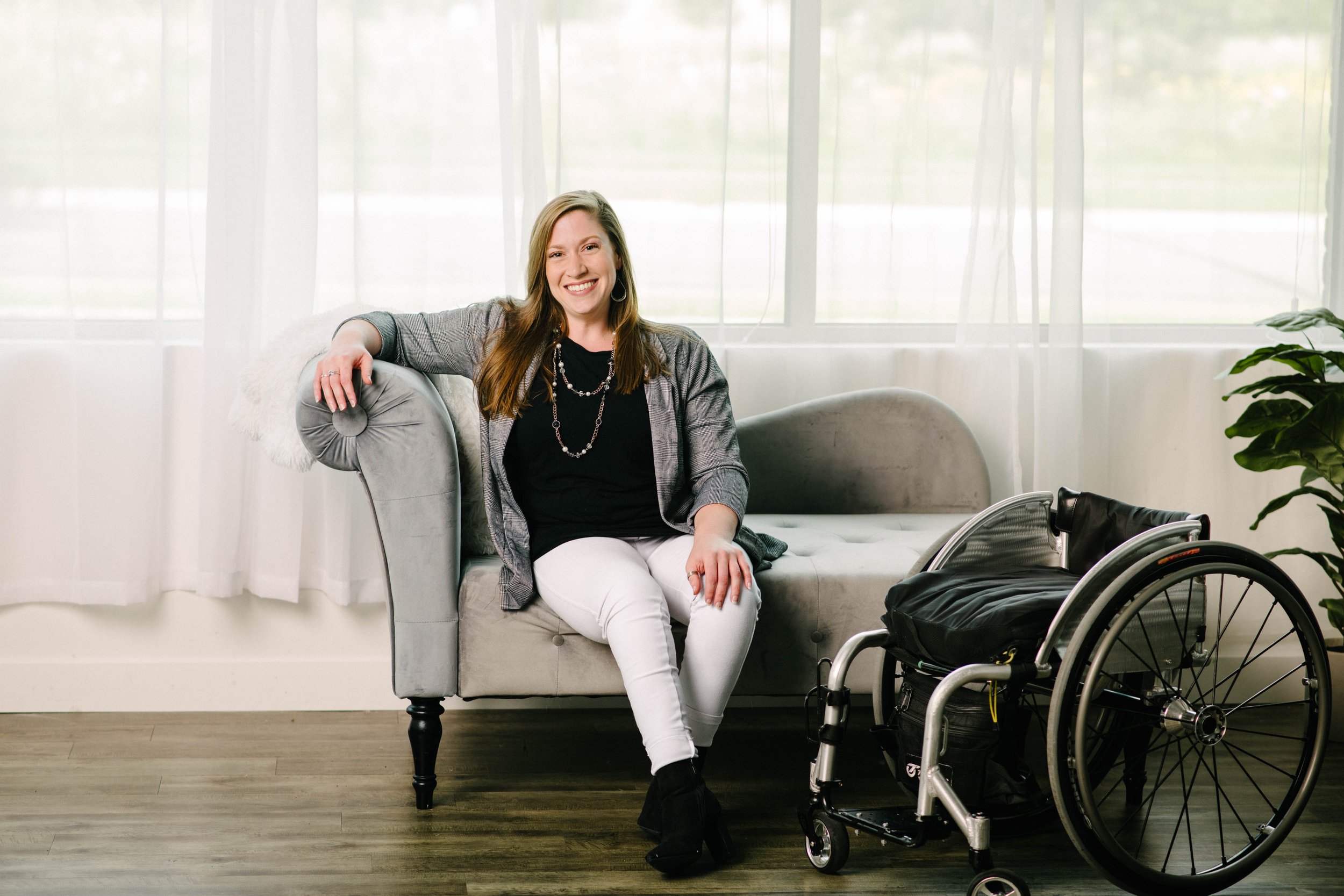 A woman with long brown hair, smiling, sitting on a light gray sofa in a bright room with white curtains and a wooden floor. There is a wheelchair positioned next to her and a potted plant on the right.